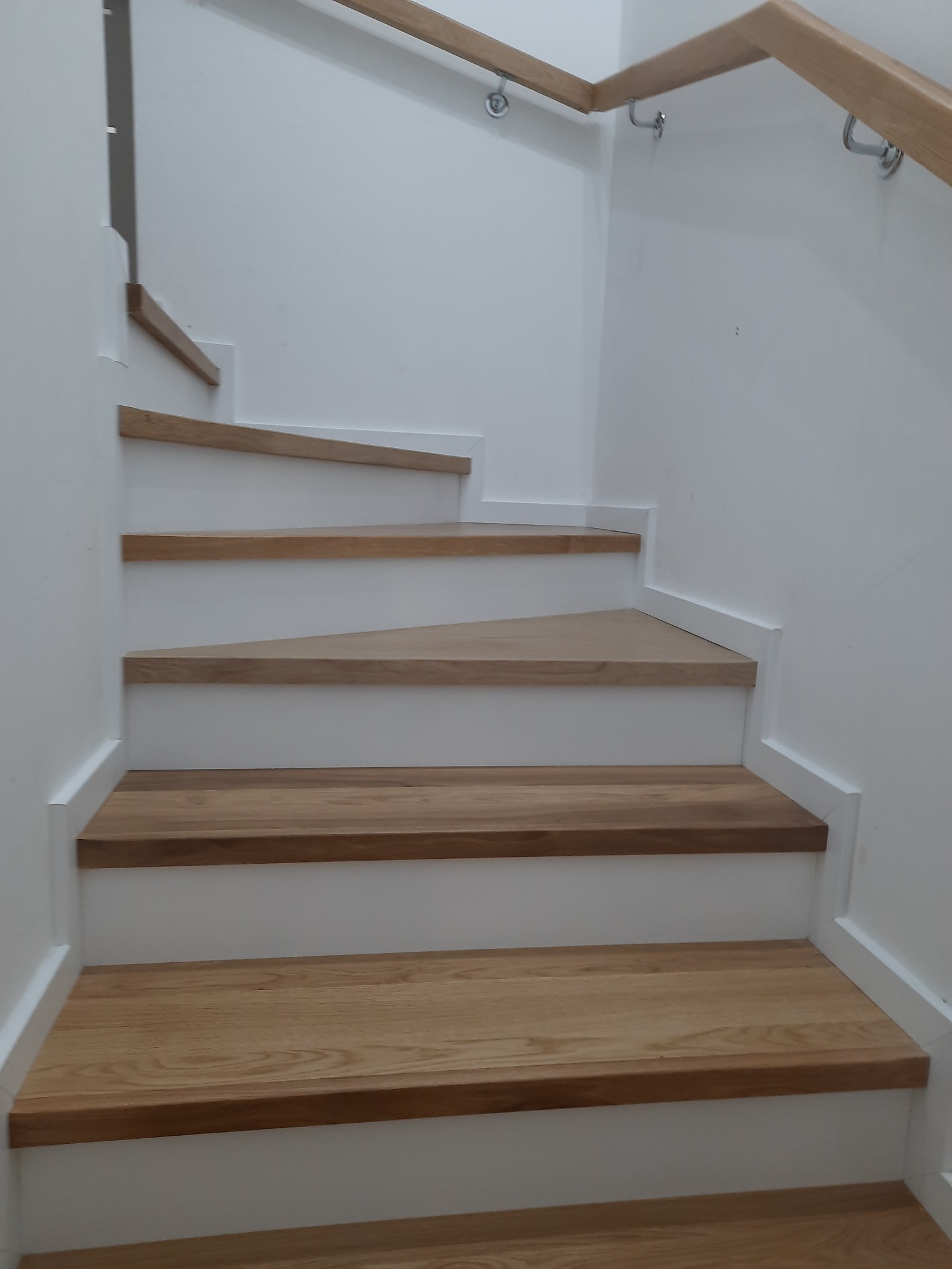 Drewniane schody z poręczą, białe stopnice, naturalny odcień drewna. Ujęcie od dołu, widoczne mocowania poręczy. Minimalistyczny design.