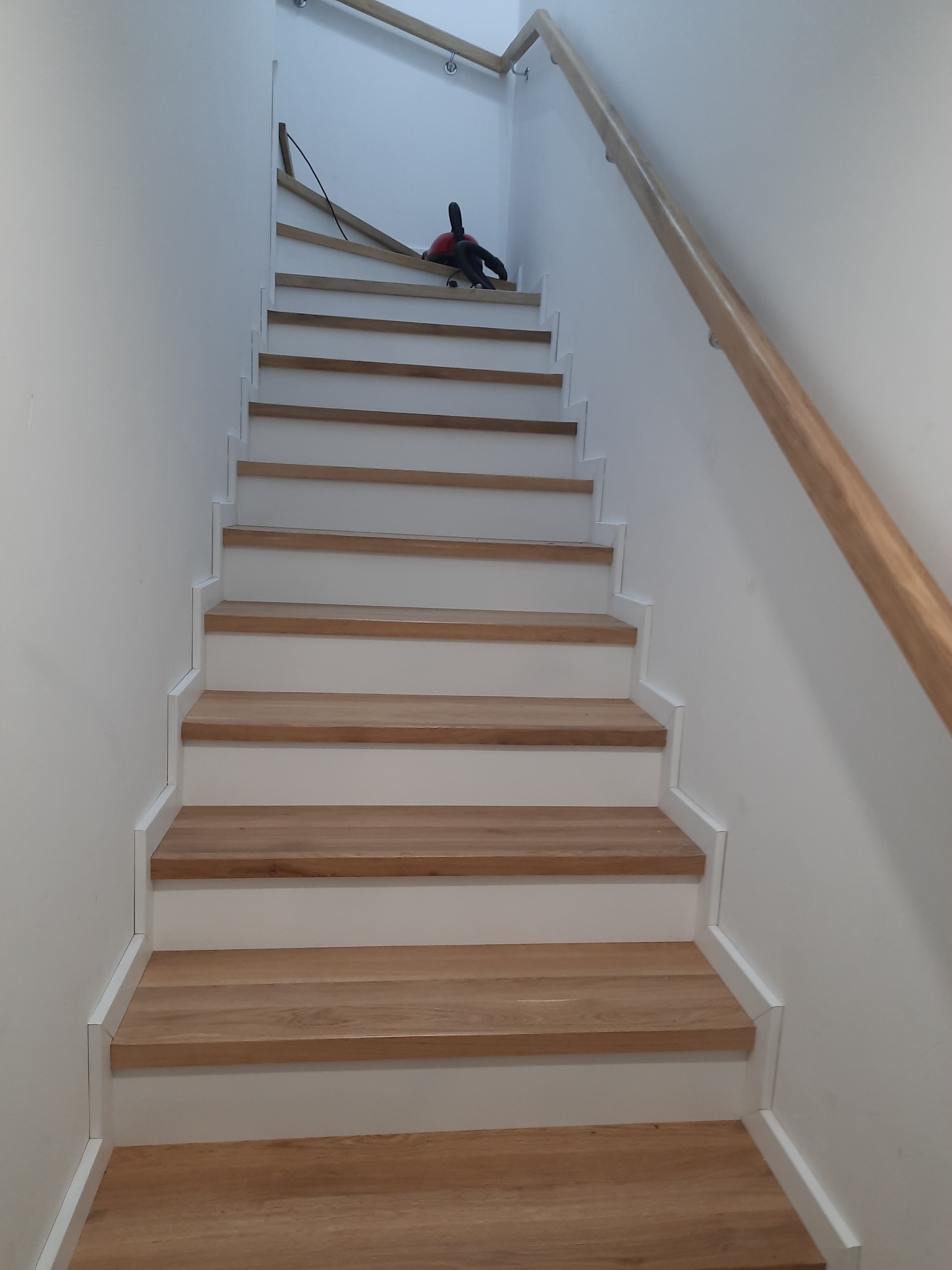 Bieg schodów z drewnianymi stopniami i białą podstopnicą, widok z dołu. Po prawej stronie ściany zamontowana drewniana poręcz. Na górze widoczny odkurzacz.