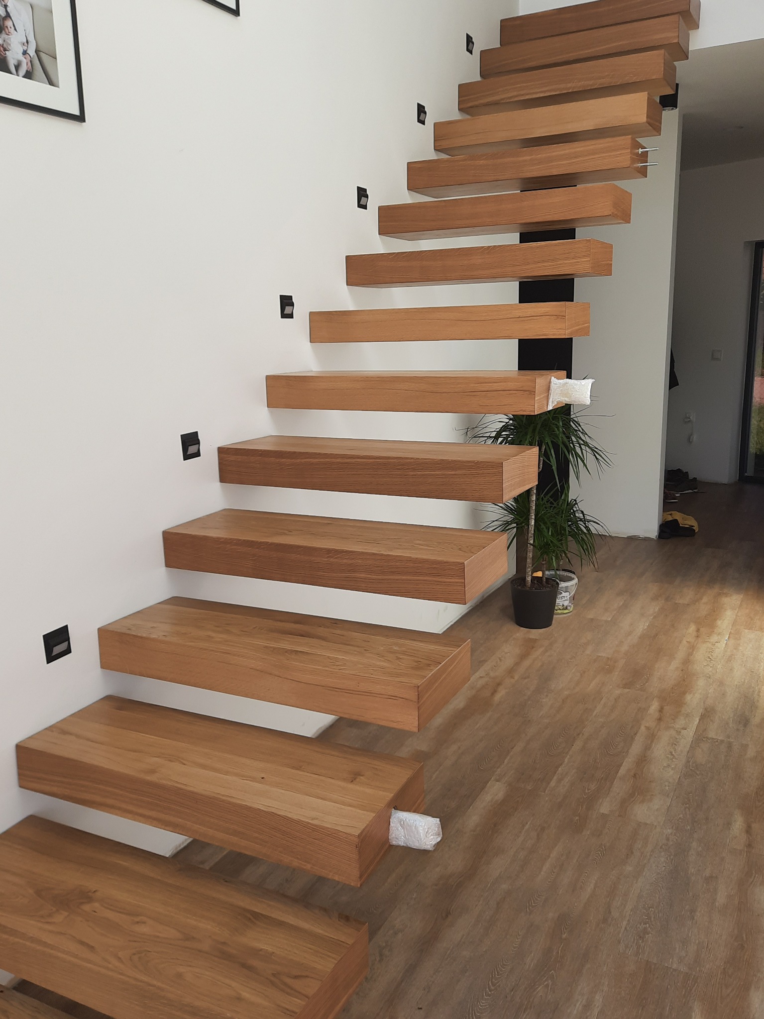 Nowoczesne, wiszące schody z drewna o ciepłym odcieniu, zamocowane do białej ściany z minimalistycznymi czarnymi punktami świetlnymi. Widok z dołu na drewnianą podłogę.