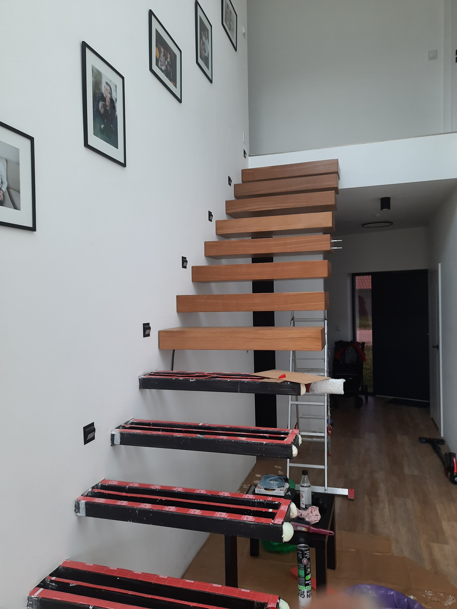 Montaż ażurowych schodów drewnianych w nowoczesnym wnętrzu. Widoczne etapy instalacji, narzędzia i zabezpieczenia. Na ścianie wiszą zdjęcia w czarnych ramkach.