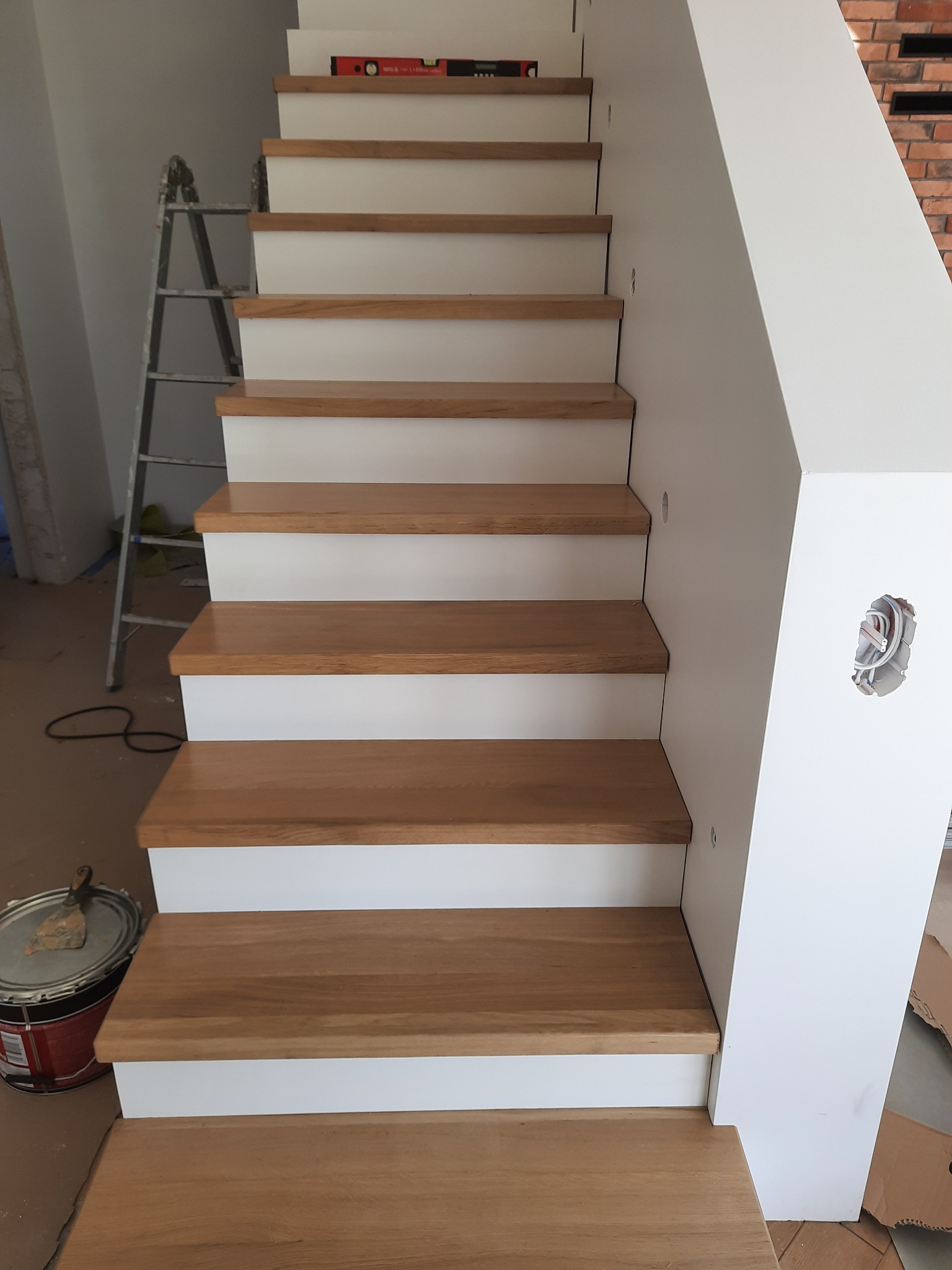 Jasne schody z drewnianymi stopniami i białymi podstopnicami w trakcie montażu, widoczna drabina i puszka elektryczna w ścianie.