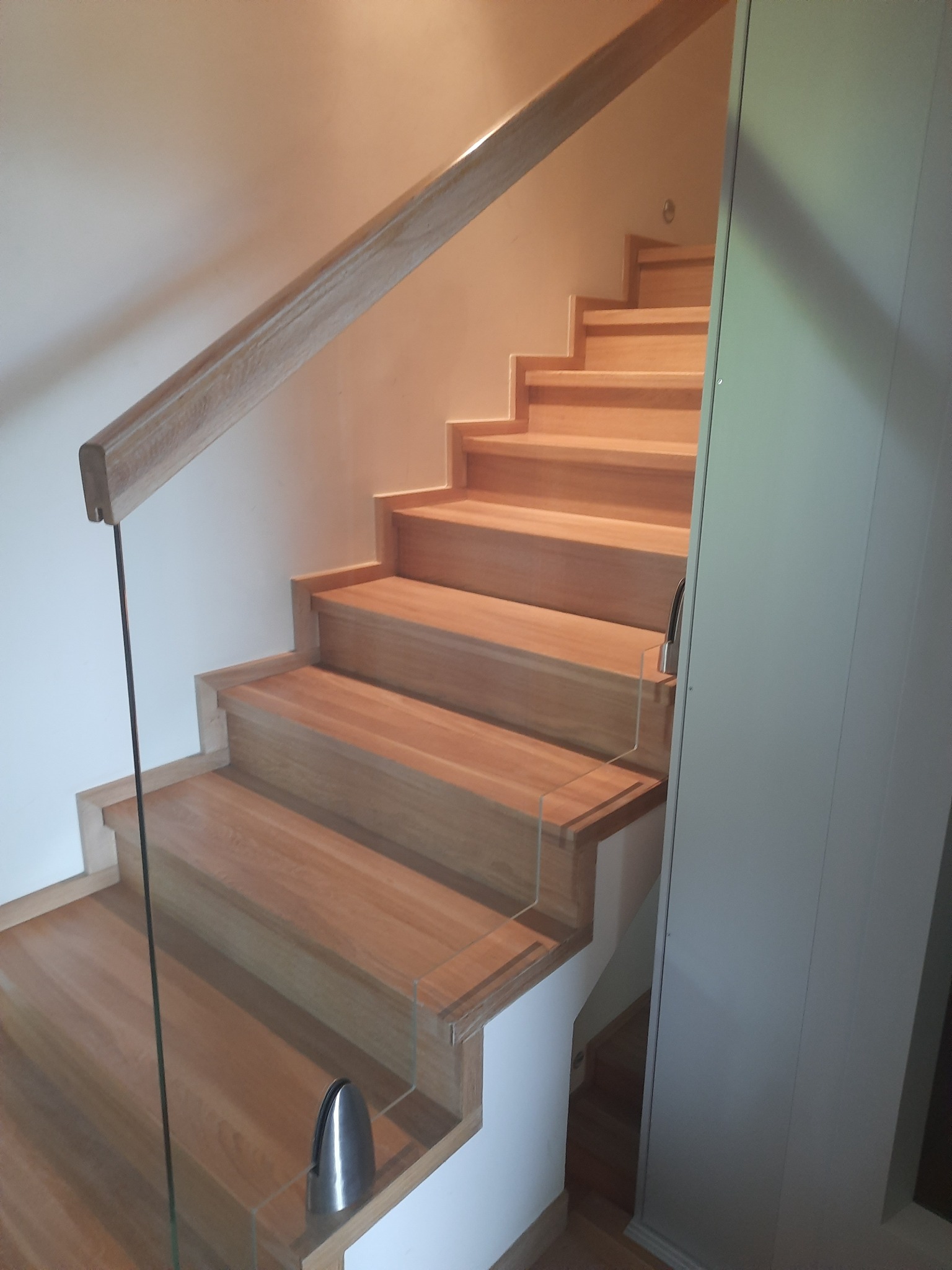 Jasne, drewniane schody z delikatną szklaną balustradą, oświetlone ciepłym światłem, prowadzące do góry. Minimalistyczny design z nowoczesnymi detalami.