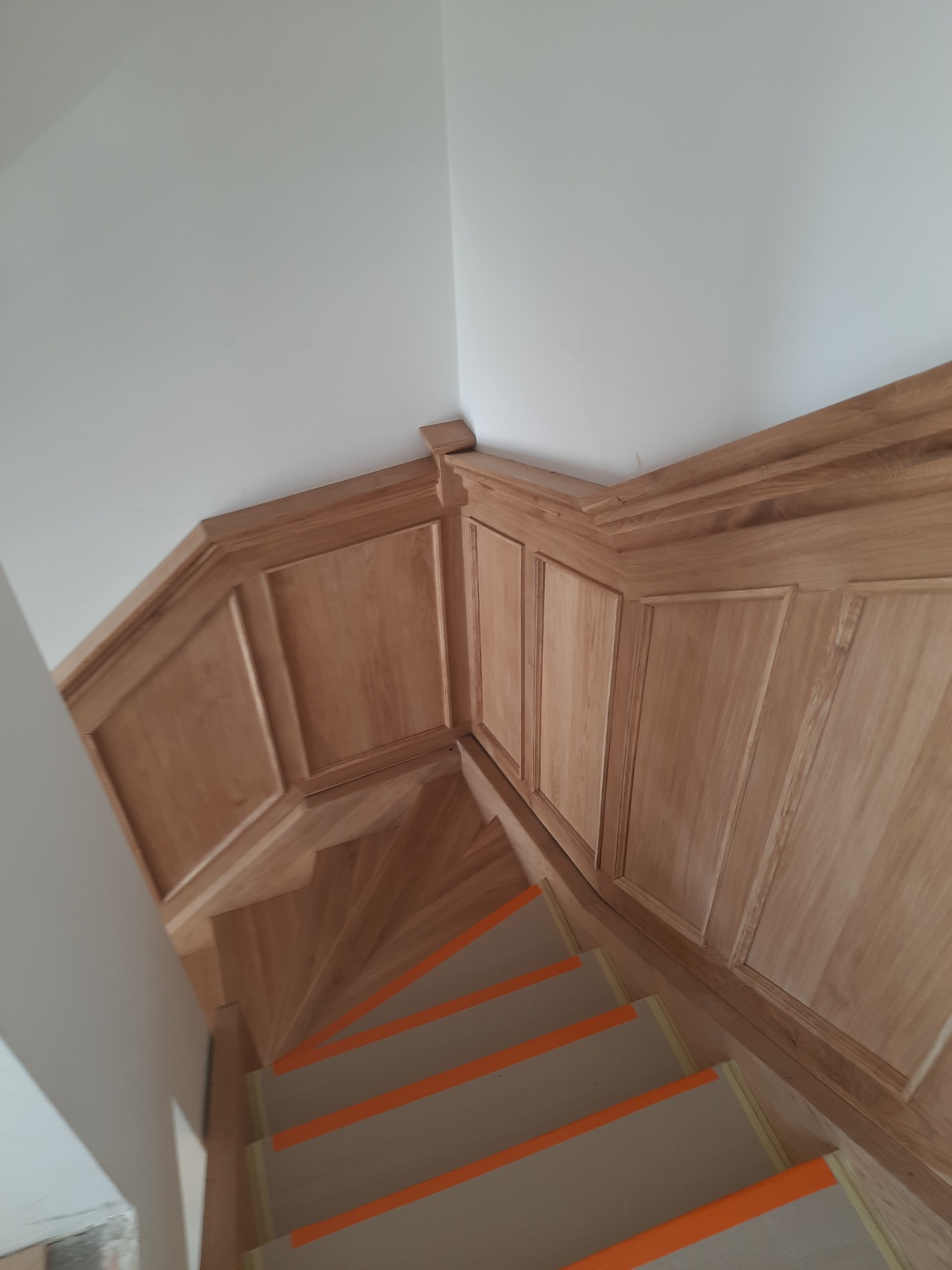 Kątowe ujęcie drewnianych schodów z panelami ściennymi. Widoczne pomarańczowe zabezpieczenia na stopniach i eleganckie wykończenie paneli przy suficie.