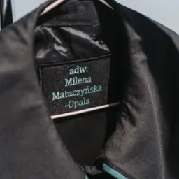 Czarna toga adwokacka z wyhaftowanym imieniem i nazwiskiem adwokat Mileny Mataczyńskiej-Opala, wisząca na różowym wieszaku na tle jasnej ściany.