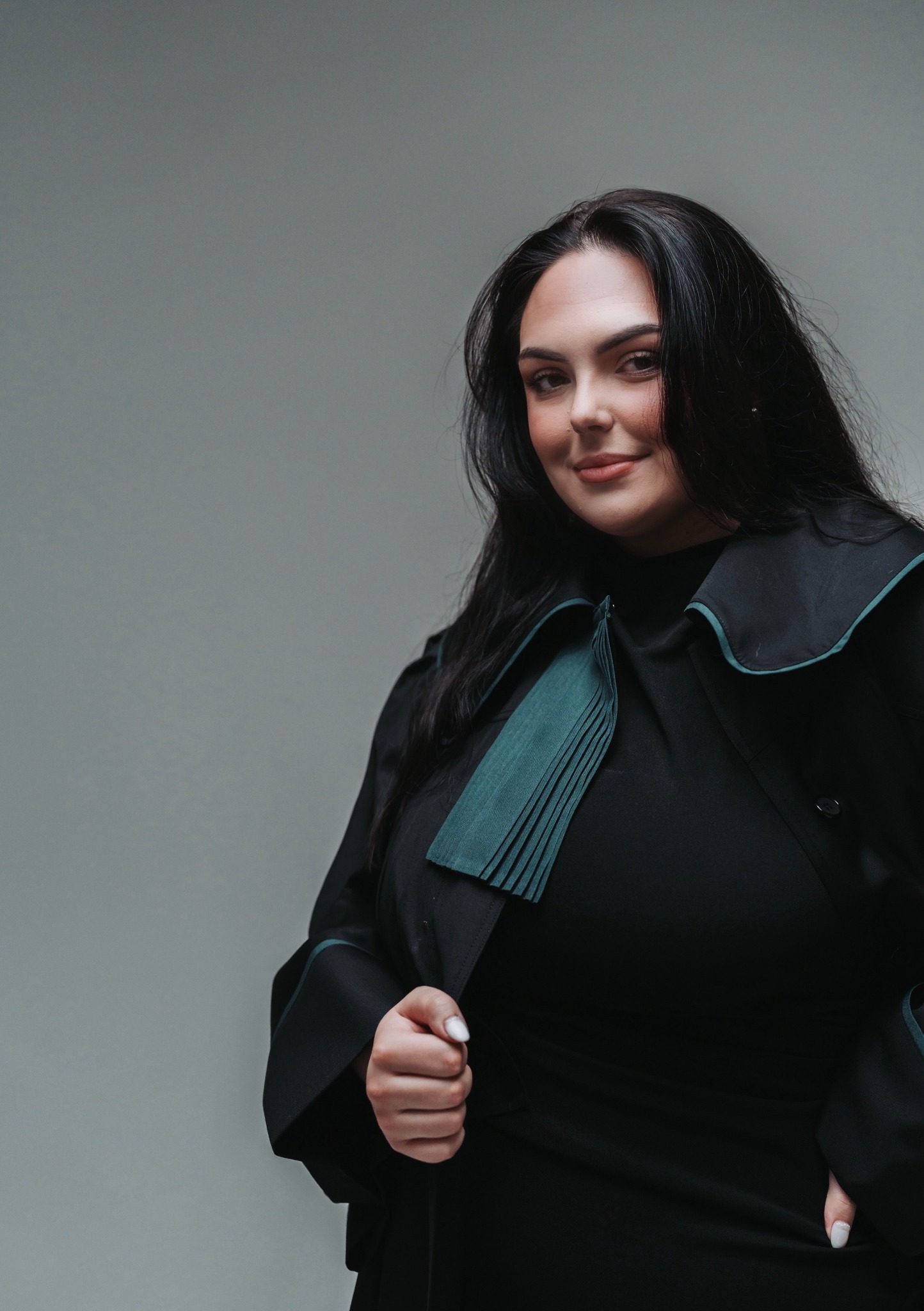 Portret kobiety-adwokata w todze z zielonym żabotem, uśmiechającej się do kamery na neutralnym tle, z ręką na klapie togi.