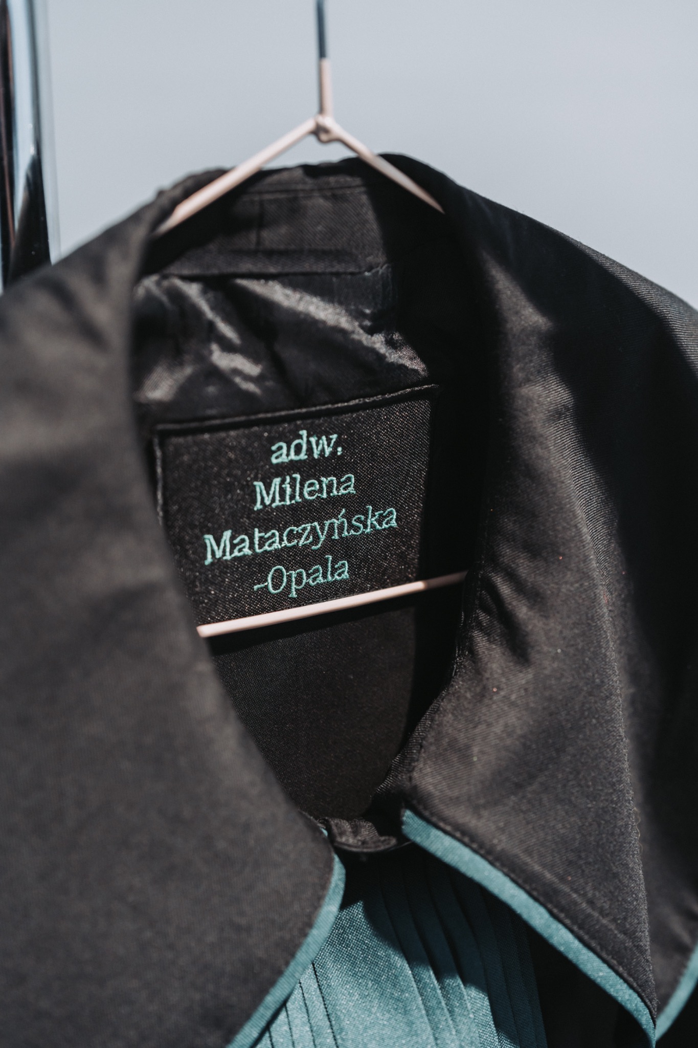 Czarna toga adwokacka z wyhaftowanym imieniem i nazwiskiem adwokat Mileny Mataczyńskiej-Opala, wisząca na różowym wieszaku na tle jasnej ściany.