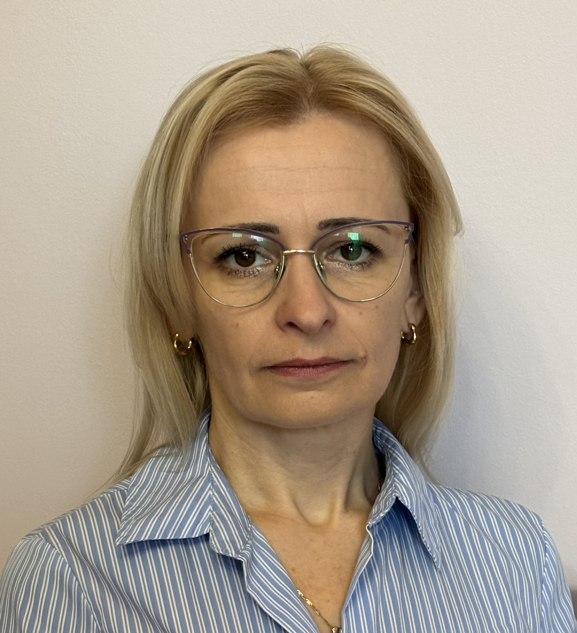 Portret kobiety w okularach i błękitnej koszuli w paski, z blond włosami i złotą biżuterią, na jasnym tle. Profesjonalny wizerunek.