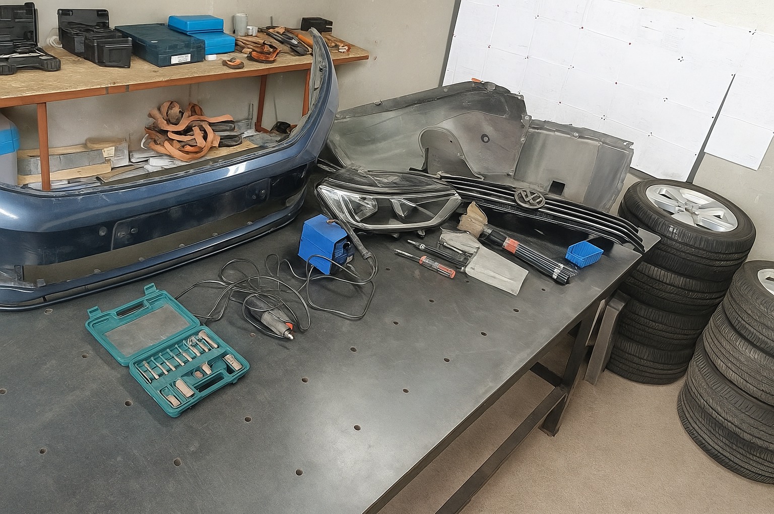 Warsztat samochodowy: zderzak, lampa, grill VW i opony na stole warsztatowym. Widoczne narzędzia i elementy karoserii. Przygotowanie do naprawy.