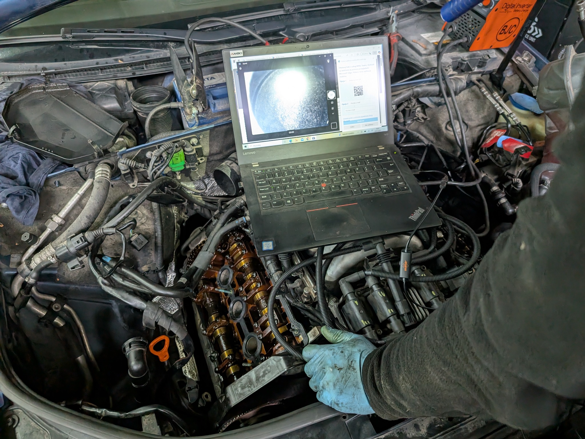 Diagnostyka silnika auta z użyciem endoskopu i laptopa. Widoczny otwarty silnik i ekran laptopa z obrazem z kamery. Ręka mechanika w rękawicy. Specjalistyczny sprzęt.