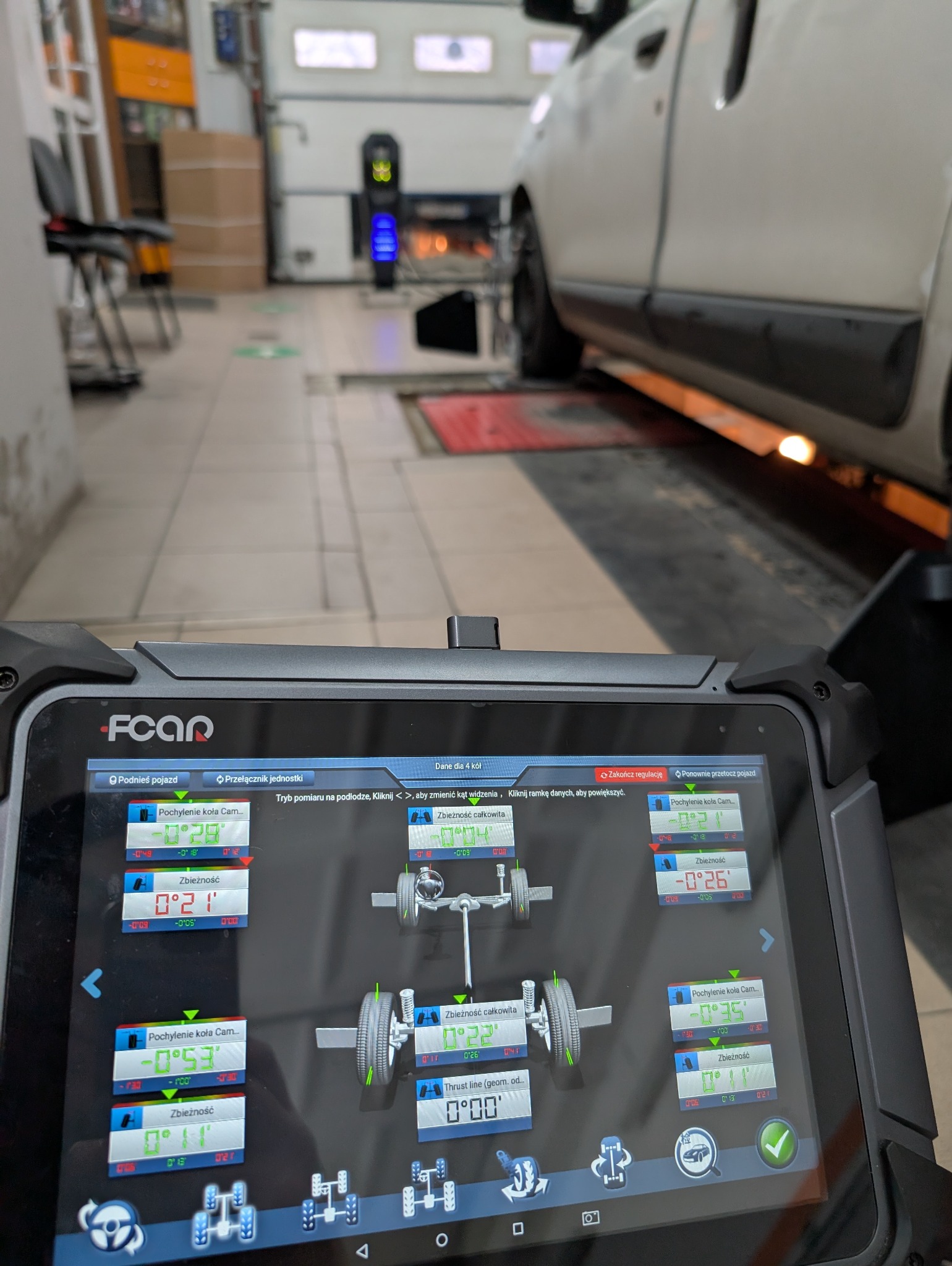 Ekran urządzenia diagnostycznego z danymi geometrii kół samochodu w warsztacie. Widoczny fragment pojazdu na stanowisku pomiarowym i narzędzia w tle.