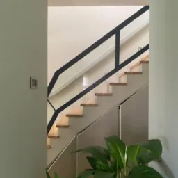 Schody z drewnianymi stopniami i czarną balustradą z elementami szkła, widoczne zza fragmentu ściany, z rośliną doniczkową w dolnej części kadru.