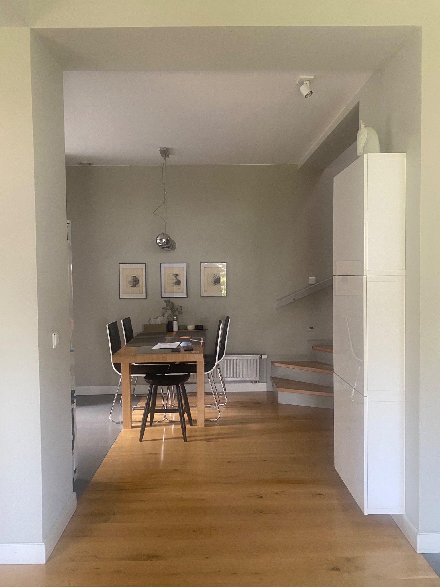 Nowoczesna jadalnia z drewnianą podłogą, stołem z krzesłami i minimalistycznymi dekoracjami ściennymi. Biała szafa i schody z drewna w tle.