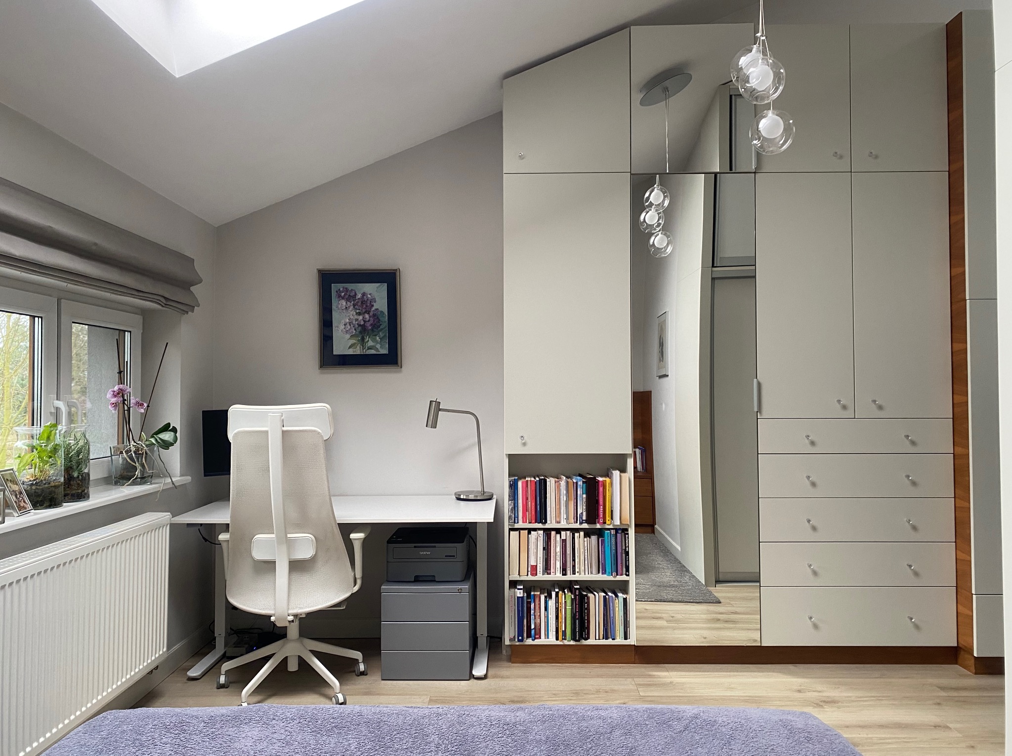 Jasne biuro w domu: białe biurko z krzesłem, regał z książkami i szafa z lustrem. Skos dachu z oknem dodaje naturalnego światła. Minimalistyczny design.