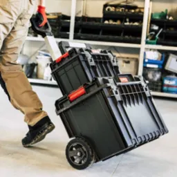 Mobilny warsztat: Czarny zestaw skrzynek narzędziowych na kółkach, ciągnięty przez osobę w roboczych spodniach i rękawicach na tle regału warsztatowego.