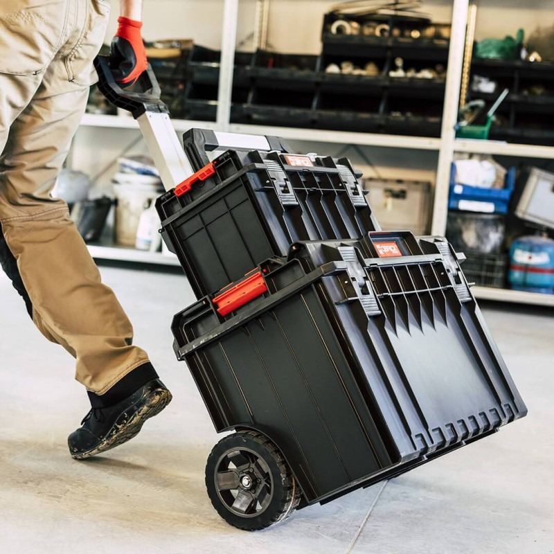 Mobilny warsztat: Czarny zestaw skrzynek narzędziowych na kółkach, ciągnięty przez osobę w roboczych spodniach i rękawicach na tle regału warsztatowego.