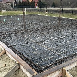 MM POLBUD Paweł Olbryś - Zbrojenie fundamentu domu jednorodzinnego z deskowaniem, widok z góry. Na drugim planie hałda piasku i zabudowa jednorodzinna.