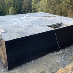 MM POLBUD Paweł Olbryś - Betonowy fundament z czarną izolacją, widok z góry, teren budowy z mikserem i drabinką, drzewa w tle.