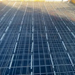 Siatka zbrojeniowa na czarnej folii, z białymi elementami dystansowymi, przygotowana do zalania betonem. Widok z góry, w kadrze widoczne deski szalunkowe.