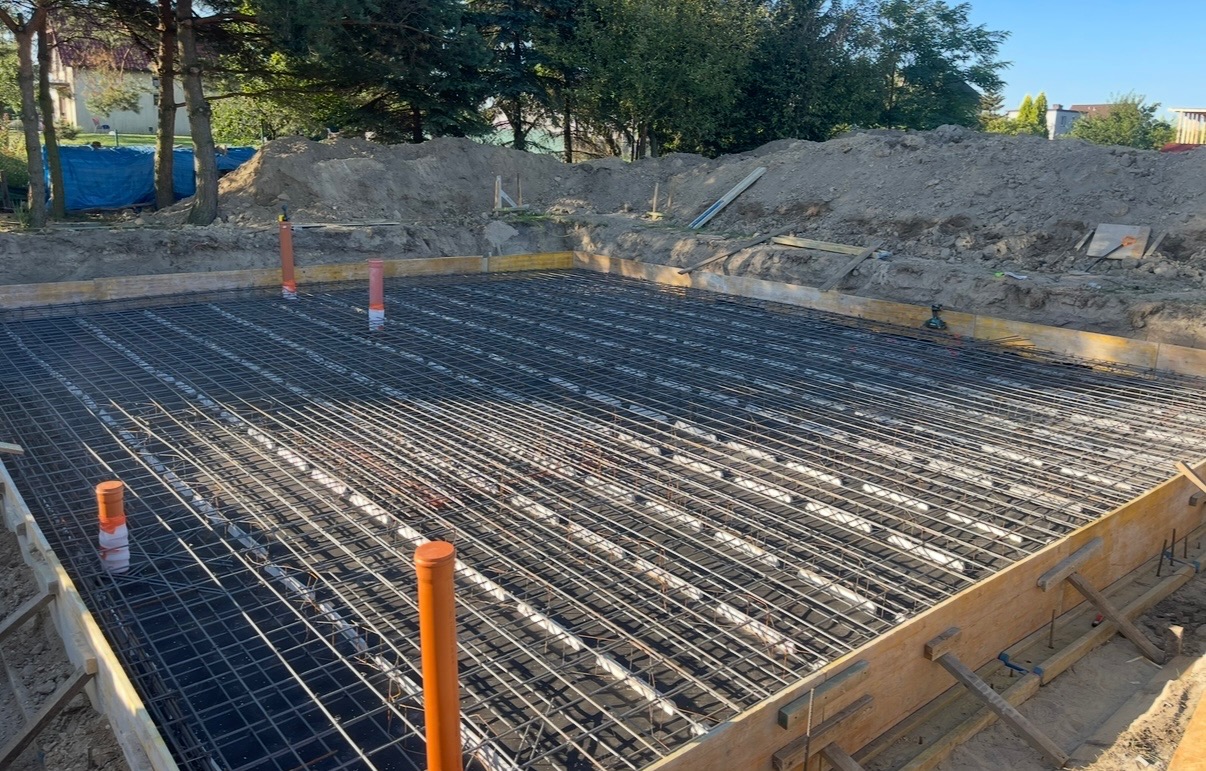 Przygotowany szalunek zbrojeniowy fundamentu pod budowę domu, widoczne pomarańczowe rury instalacyjne, w tle ziemia i drzewa.