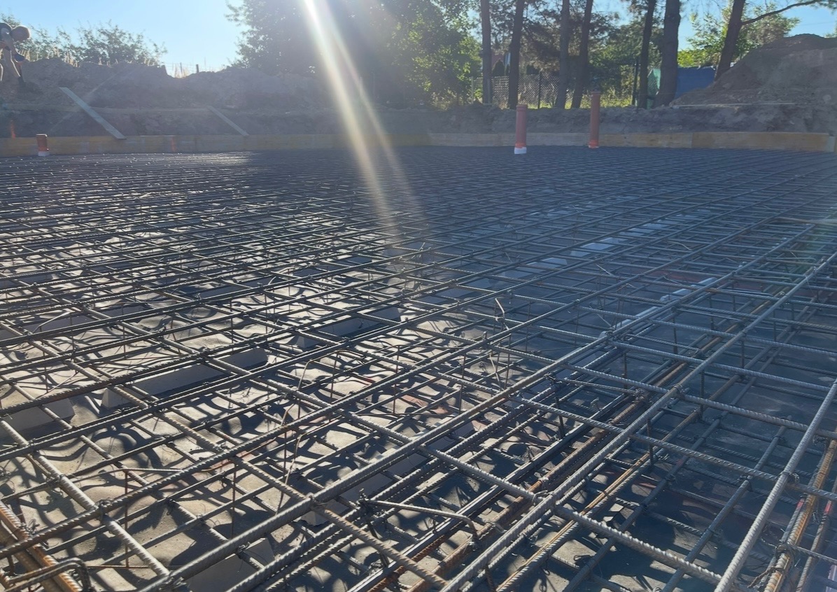 Zbrojenie fundamentu na podkładzie betonowym, widoczne rury instalacyjne i szalunek. Jasne światło słoneczne podkreśla detale konstrukcji.