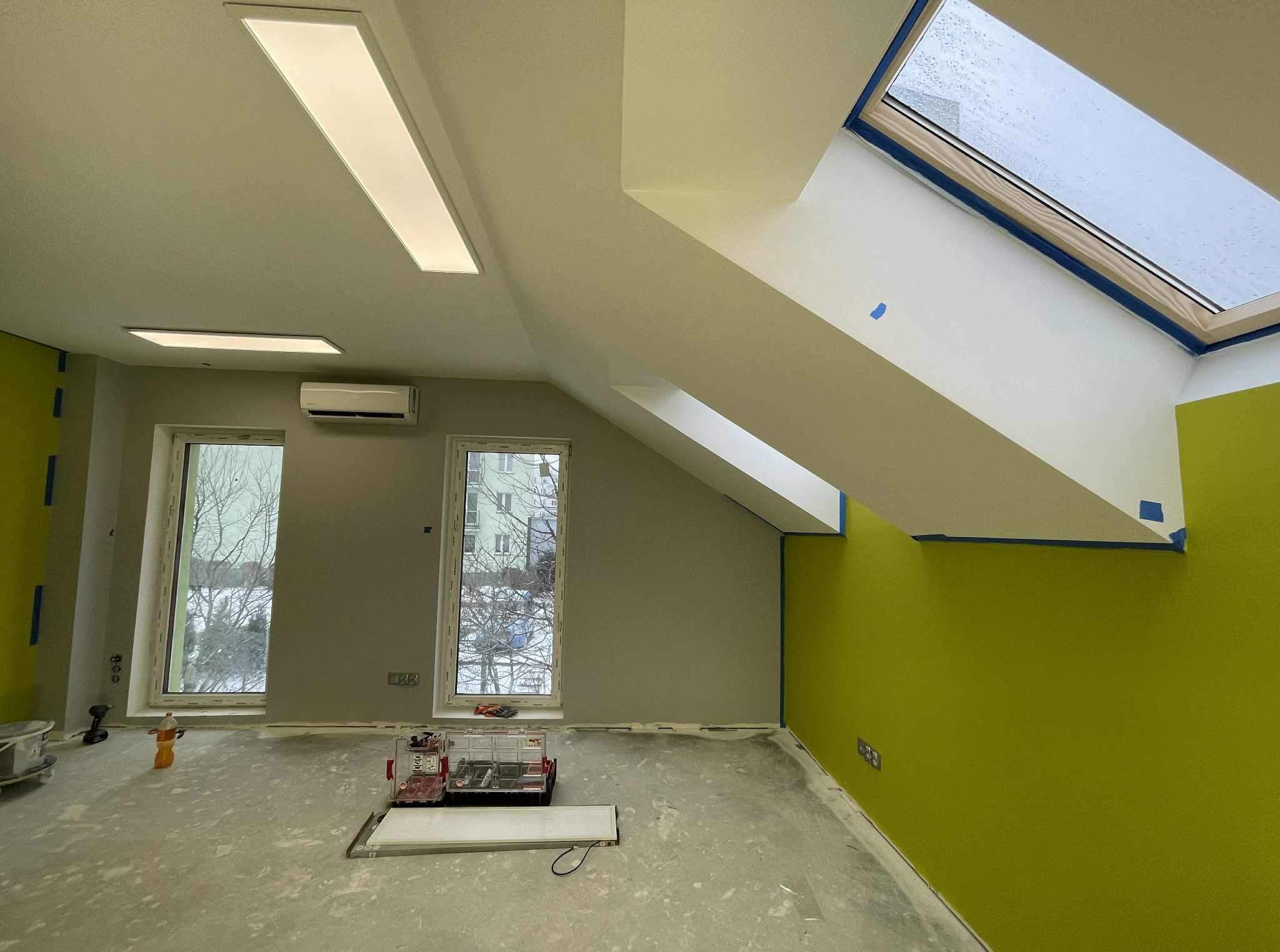 Poddasze po malowaniu: zielona ściana kontrastuje z szarą. Widoczne okna dachowe z niebieską taśmą malarską, grzejnik i narzędzia malarskie. Powierzchnia podłogi zabezpieczona.