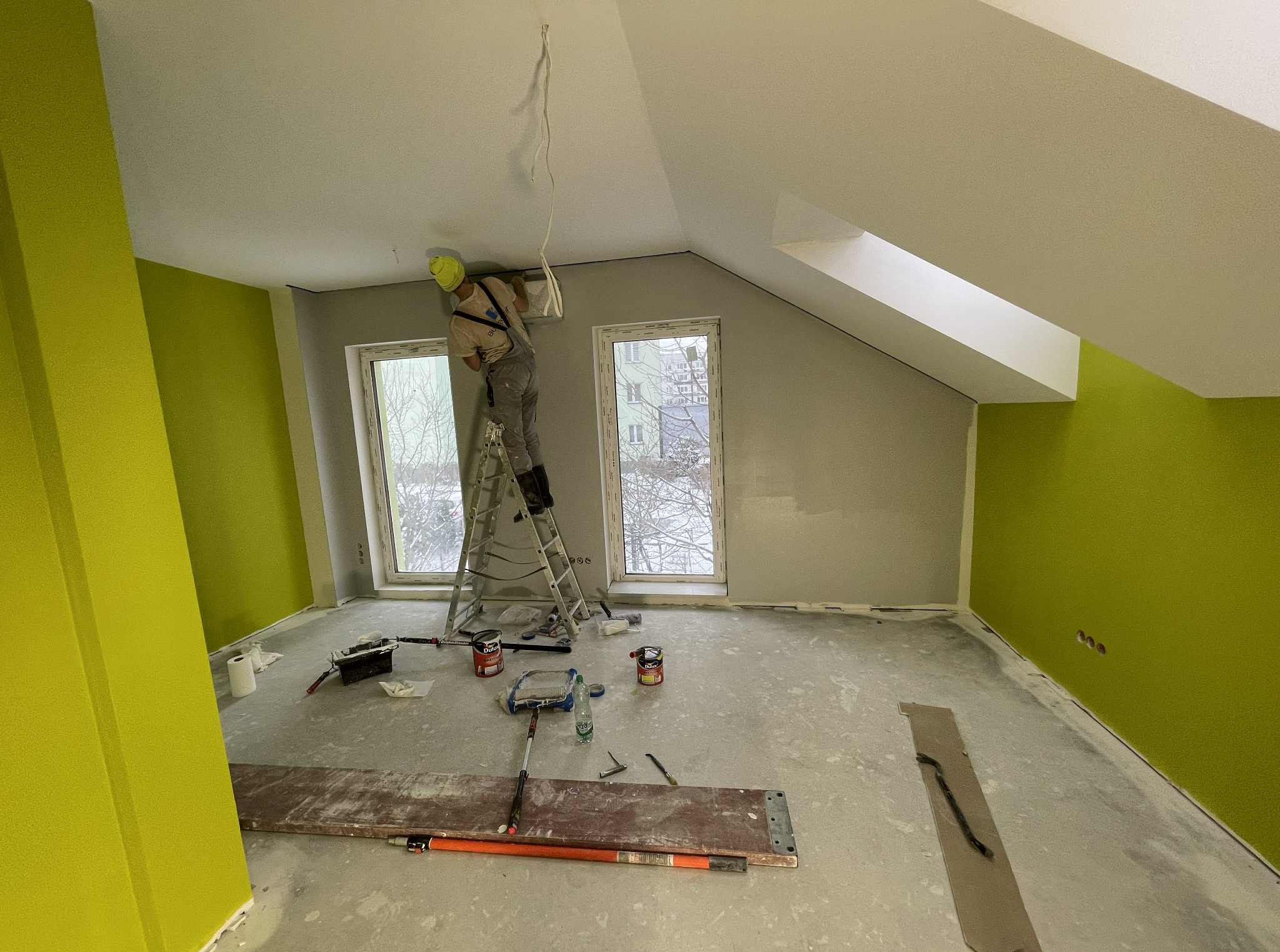 Malowanie poddasza w Gliwicach: malarz na drabinie, ściany w kolorze limonki i szarości, widoczne narzędzia malarskie i okno dachowe. Prace wykończeniowe.