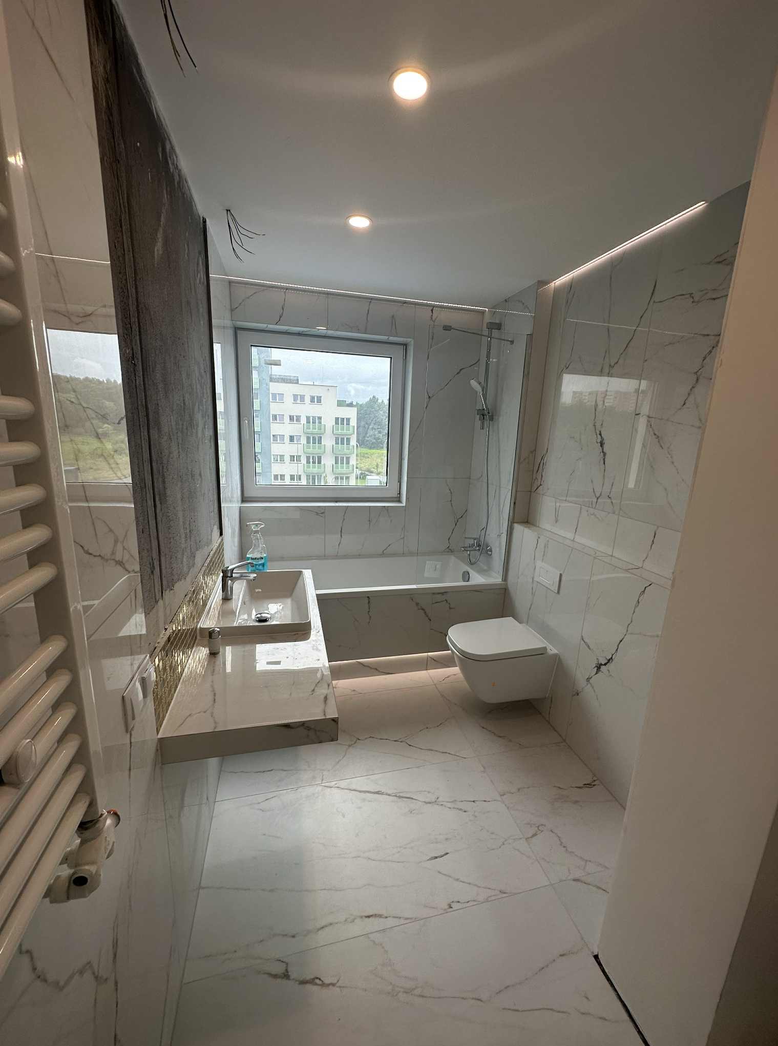 Wykończona łazienka z marmurowymi płytkami, podświetleniem LED i oknem z widokiem na budynek. Umywalka z baterią i lustrem, toaleta podwieszana, wanna z parawanem.