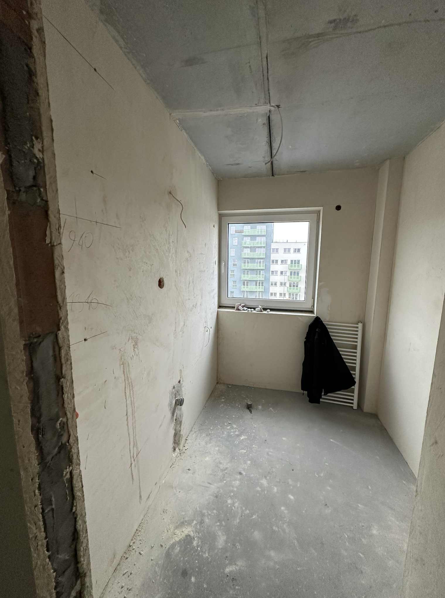 Surowy pokój w trakcie remontu: szpachlowane ściany z widocznymi śladami po instalacjach, okno z grzejnikiem i wiszącą kurtką. Widoczne niedokończone wykończenia.