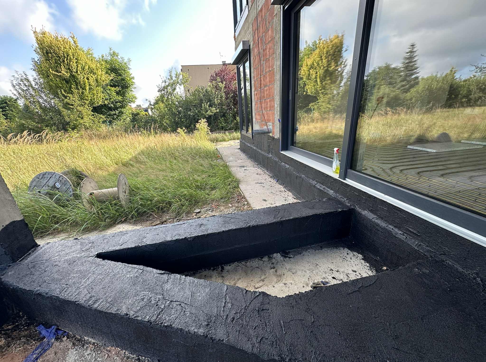 Hydroizolacja fundamentów domu w trakcie prac. Widoczne czarne zabezpieczenie przeciwwilgociowe na betonowej konstrukcji. W tle trawa i budynek.
