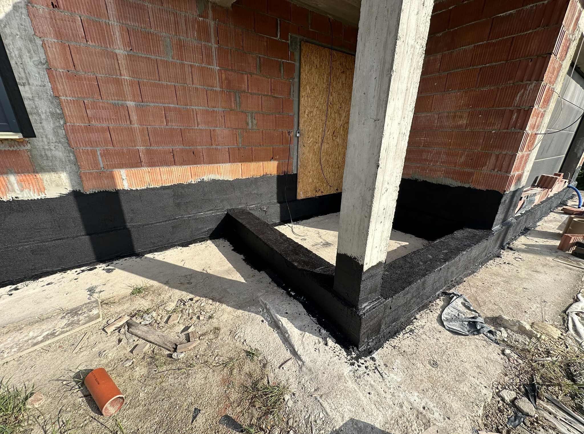 Hydroizolacja fundamentów domu z czerwonej cegły, zabezpieczona czarną powłoką bitumiczną, widok z poziomu gruntu, betonowy filar w kadrze.