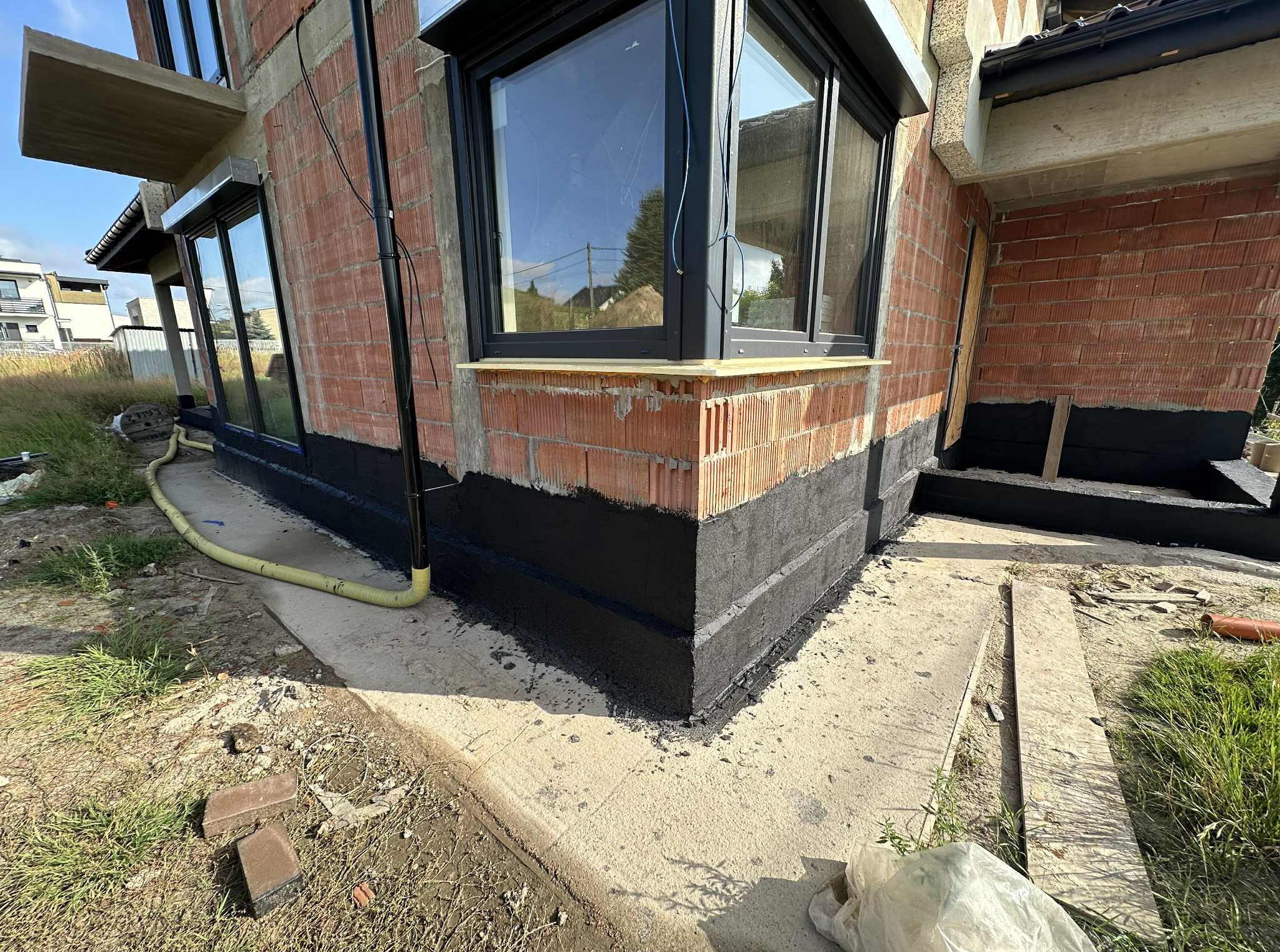 Fragment domu w trakcie prac izolacyjnych fundamentów. Widoczne czarne zabezpieczenie na ścianie z cegły, okno, fragment elewacji i otoczenie placu budowy.