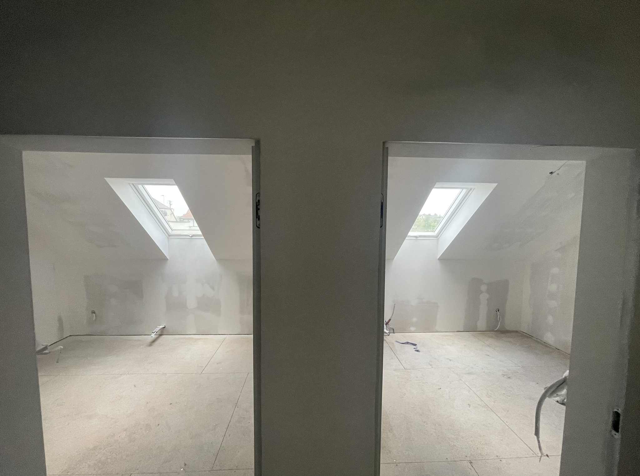 Dwa pokoje na poddaszu, widoczne przez otwory drzwiowe, ściany przygotowane do malowania, okna dachowe w skosach, surowa podłoga, widok z korytarza.