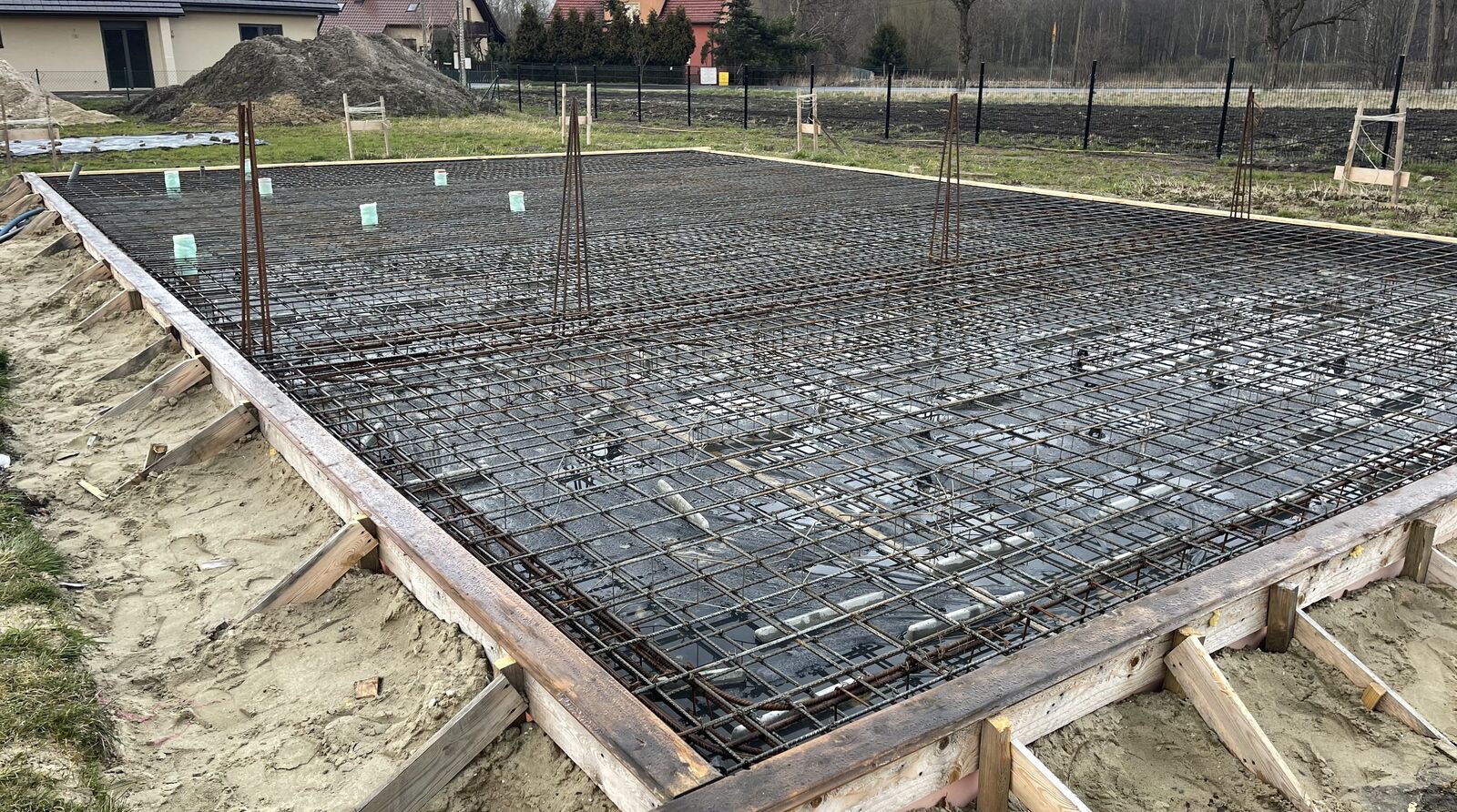 Zbrojenie fundamentu domu jednorodzinnego z deskowaniem, widok z góry. Na drugim planie hałda piasku i zabudowa jednorodzinna.