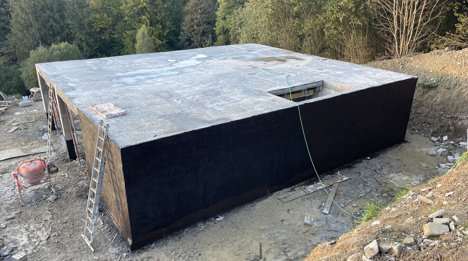 Betonowy fundament z czarną izolacją, widok z góry, teren budowy z mikserem i drabinką, drzewa w tle.