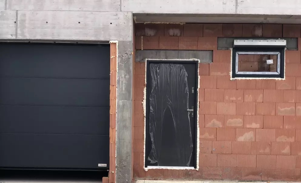 Surowy stan budynku: brama garażowa, drzwi wejściowe osłonięte folią i okno w ścianie z cegły kratówki. Widoczna pianka montażowa.