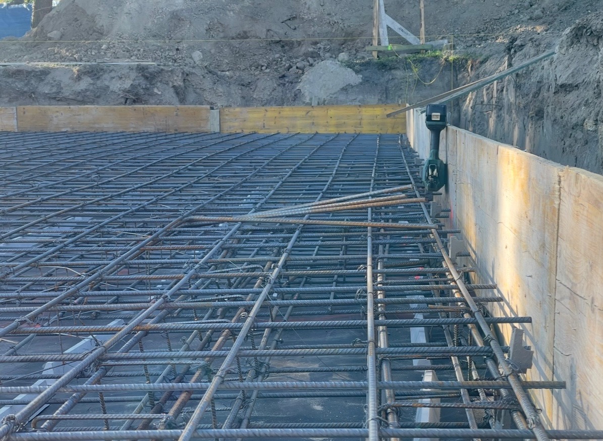 Siatka zbrojeniowa na fundamencie, widok z góry z boku. Szalunek z desek, grunt w tle. Zbrojenie wykonane z żebrowanych prętów połączonych drutem.