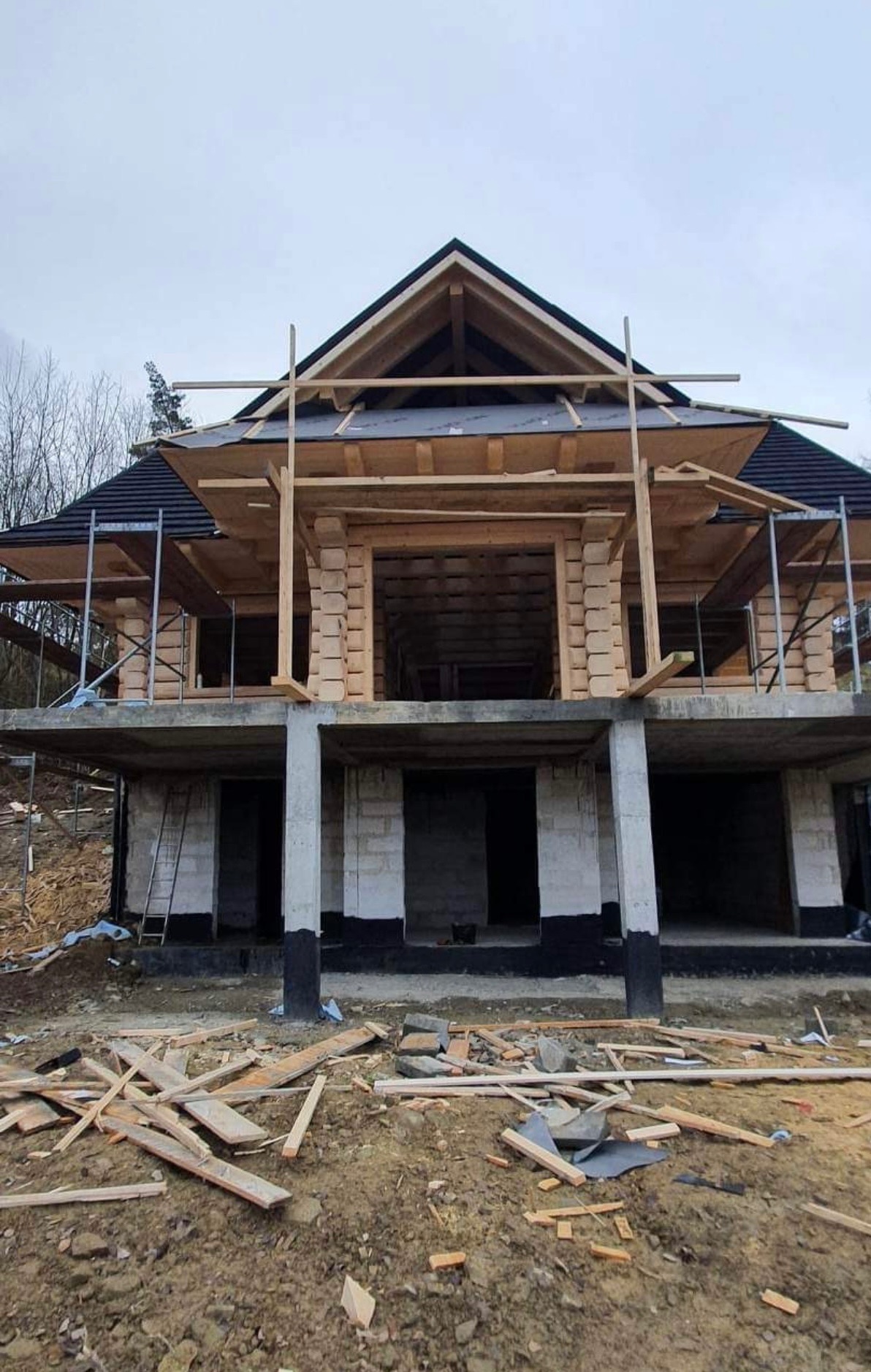 Budowa domu z bali w trakcie realizacji. Widoczna konstrukcja drewniana, fundamenty i elementy rusztowania. Na ziemi leżą odpady budowlane.