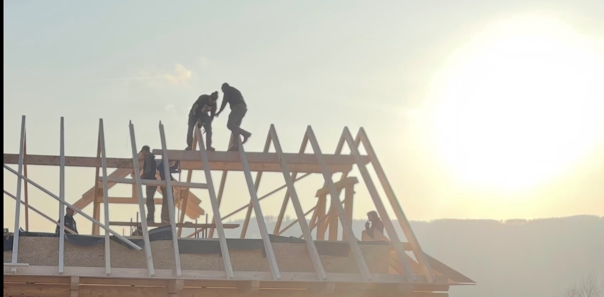 Ekipa monterów w trakcie wznoszenia drewnianej konstrukcji dachu domu jednorodzinnego na tle zachodzącego słońca. Elementy konstrukcyjne z drewna.