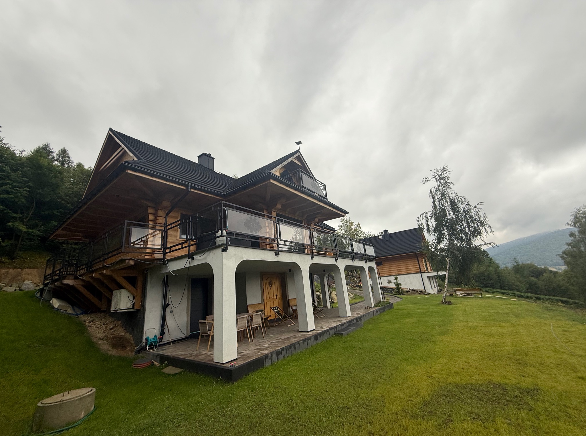 Dwukondygnacyjny dom z bali w Gliwicach, z balkonem i tarasem. Widok z trawnika, pochmurne niebo w tle. Elementy nowoczesnej architektury i naturalnego drewna.