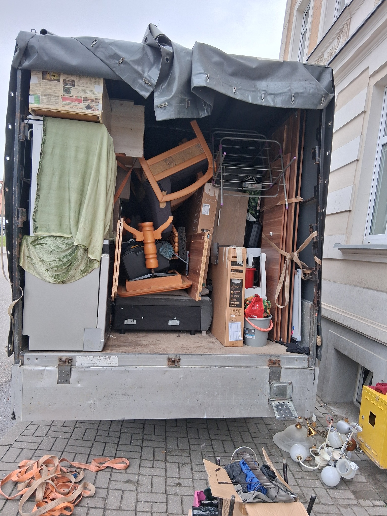 Załadunek mebli i sprzętów do ciężarówki z plandeką. Widoczne krzesła, lodówka, kartony i lampy. Część przedmiotów leży na chodniku obok pojazdu.