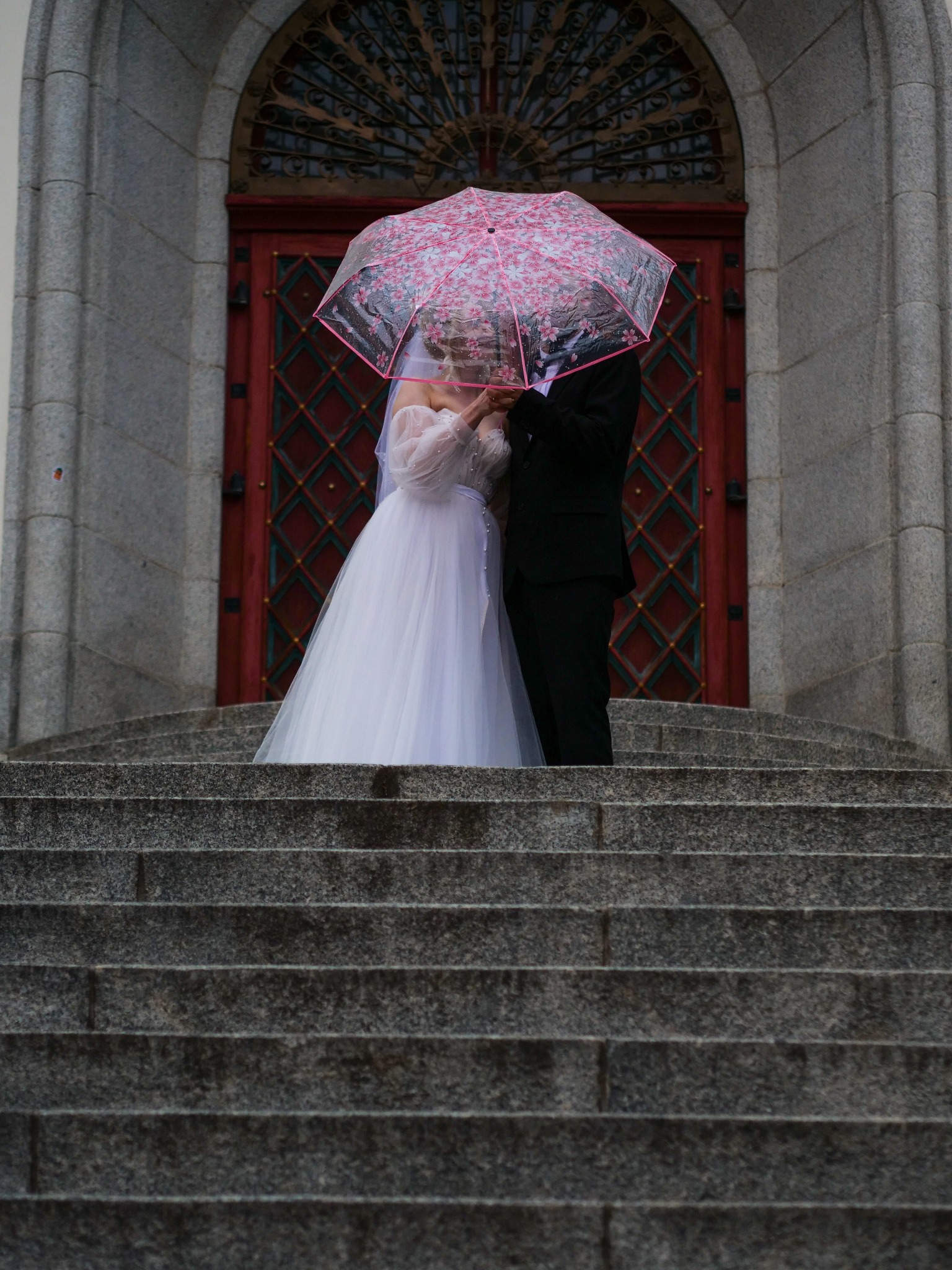 Para młoda pod transparentnym parasolem w kwiaty na schodach, na tle zdobionych drzwi. Panna młoda w białej sukni, pan młody w czarnym garniturze. Ujęcie z dołu.