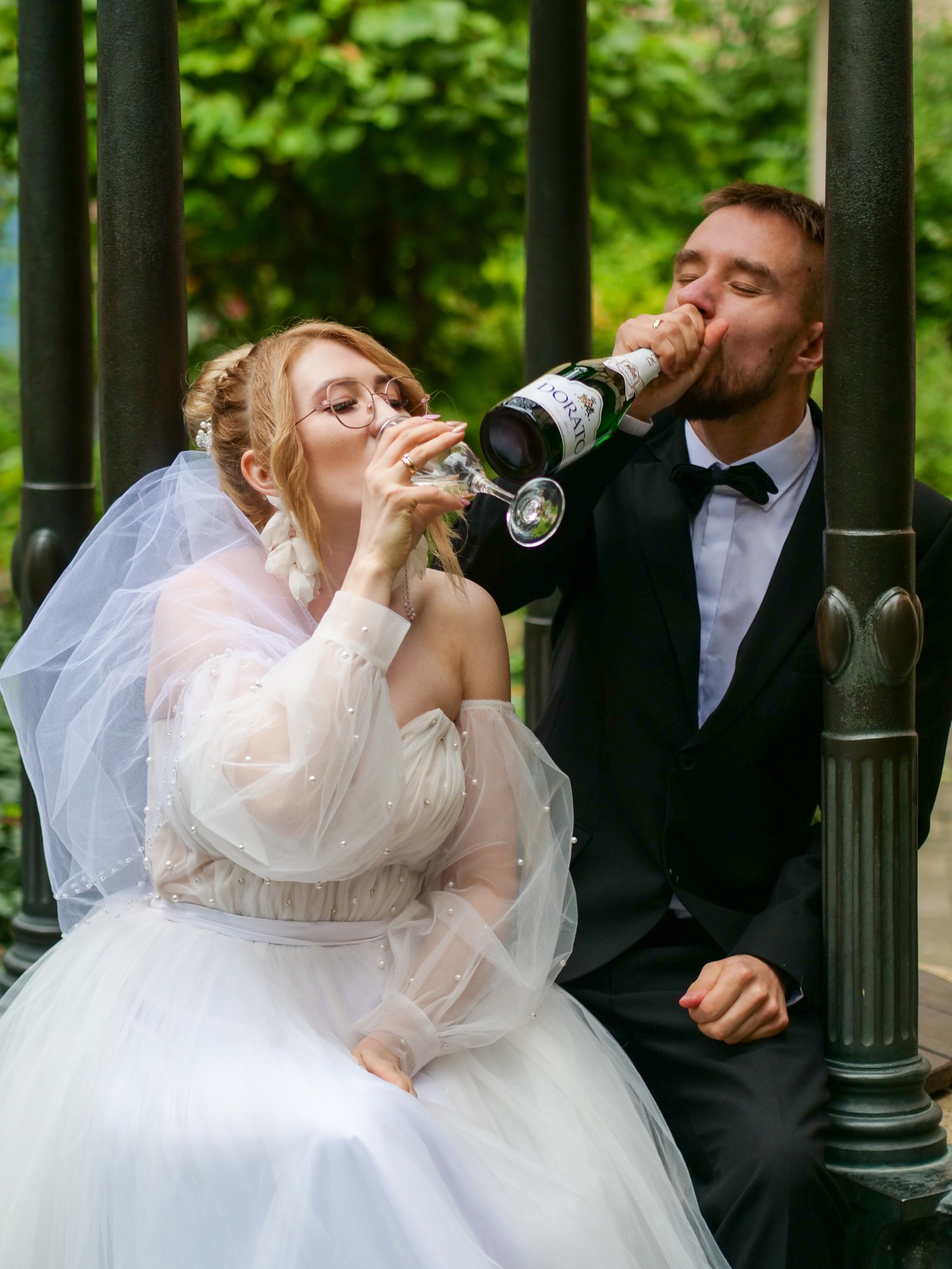 Para młoda w plenerze pije szampana: panna młoda z kieliszka, pan młody prosto z butelki Dorato. Romantyczna sesja ślubna w otoczeniu zieleni.