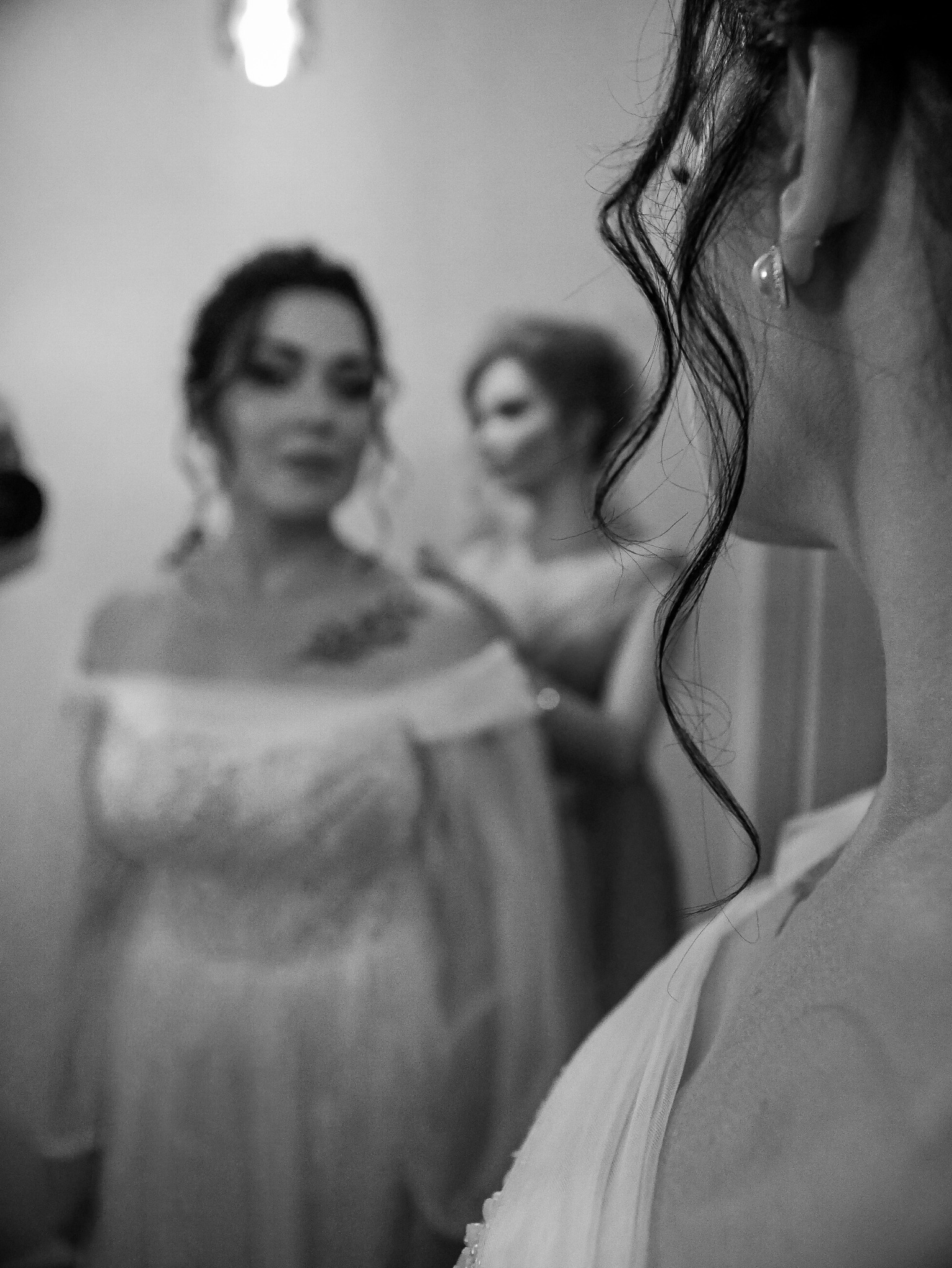 Czarno-białe zdjęcie panny młodej w sukni, z bliska widoczne kręcone włosy i kolczyk. W tle rozmyte postacie kobiety poprawiającej suknię.