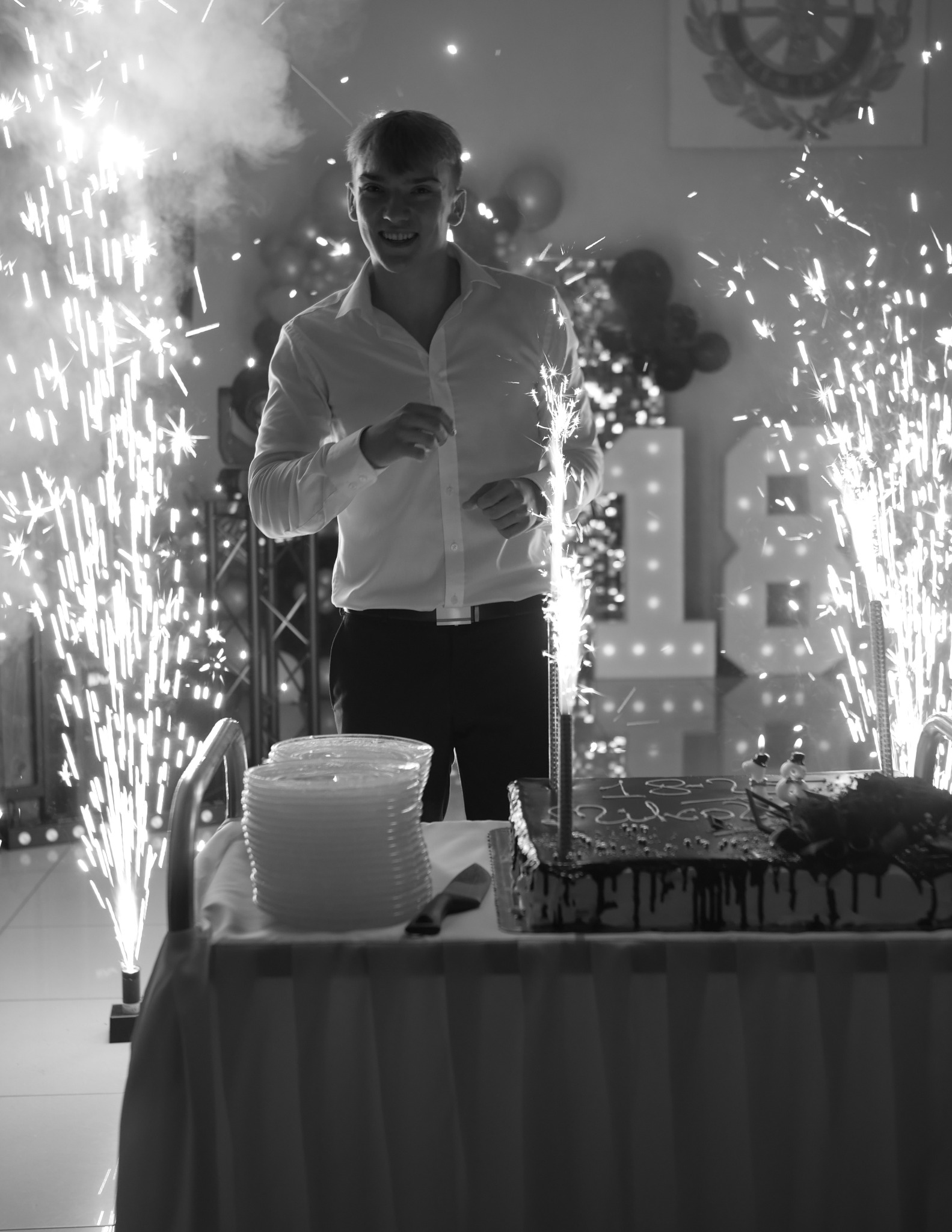 Uśmiechnięty mężczyzna w białej koszuli, tort urodzinowy z fontannami iskier w tle, dekoracje na 18 urodziny. Fotografia z Opola, czarno-biała.