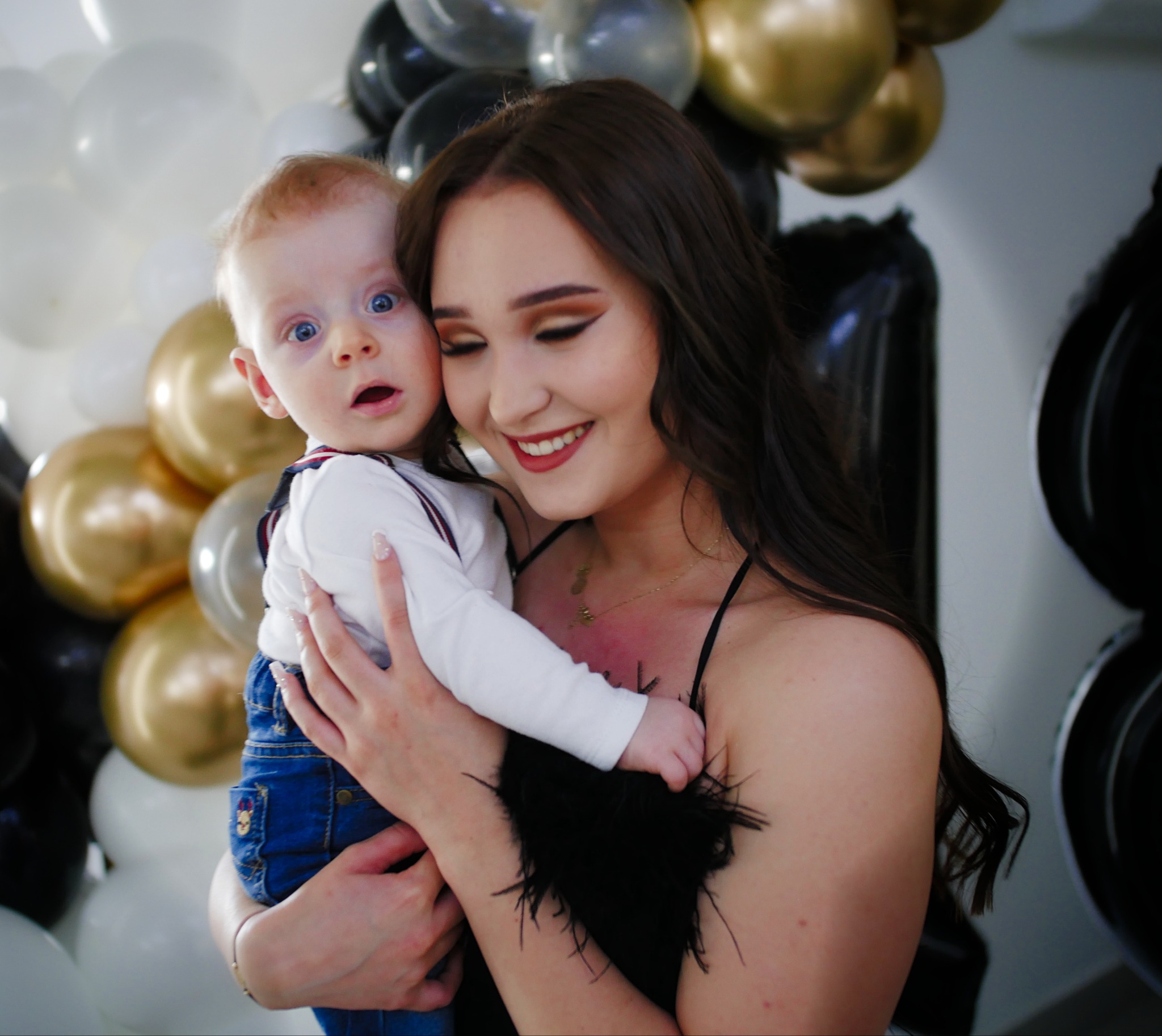 Uśmiechnięta kobieta trzyma na rękach zaskoczonego chłopca na tle dekoracji z czarno-złotych balonów. Sesja w studio fotograficznym.