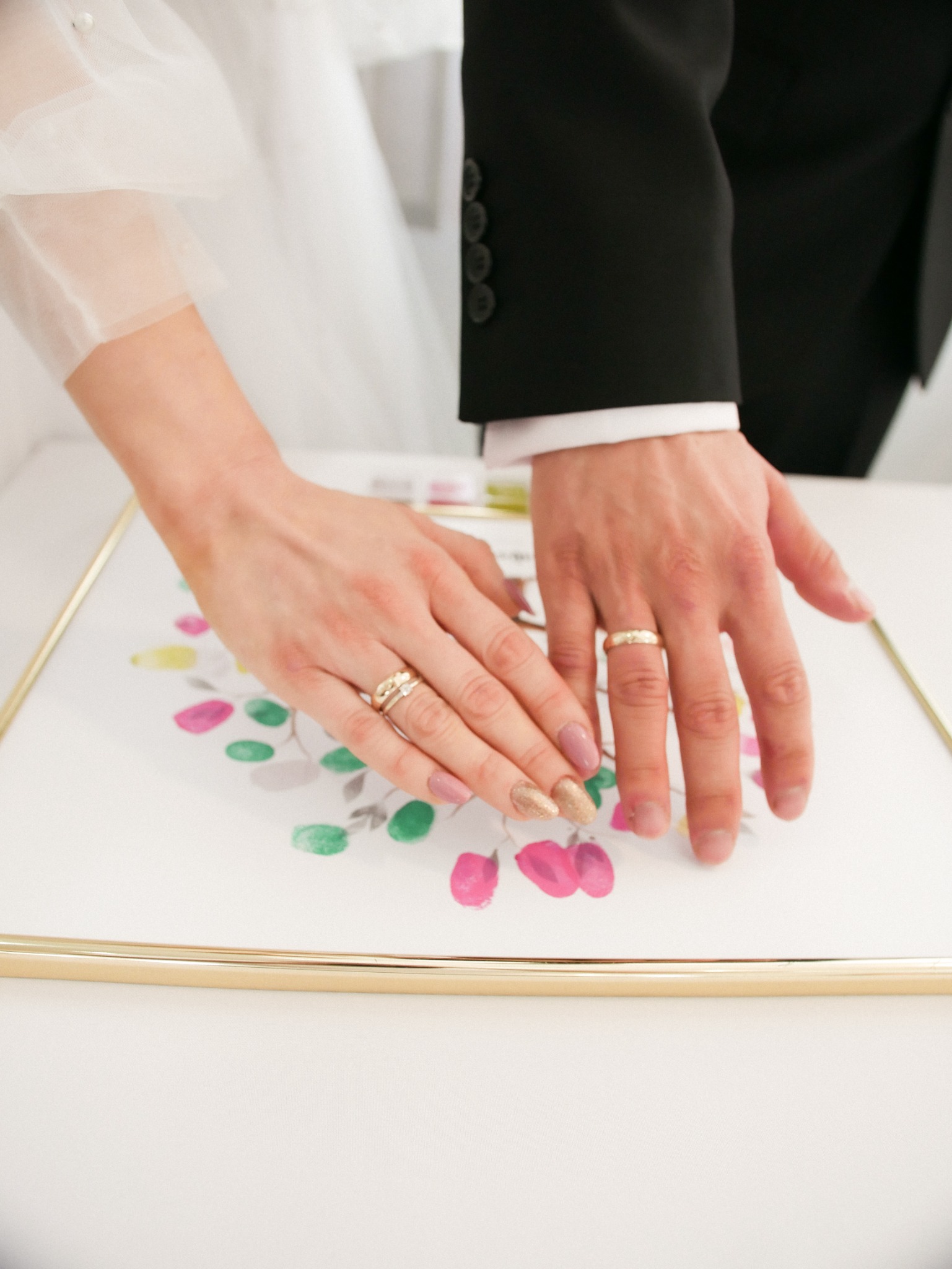 Dłonie pary młodej z obrączkami składają odciski palców na pamiątkowej grafice z motywem kwiatowym w złotej ramce. Widoczny fragment sukni ślubnej i garnituru.