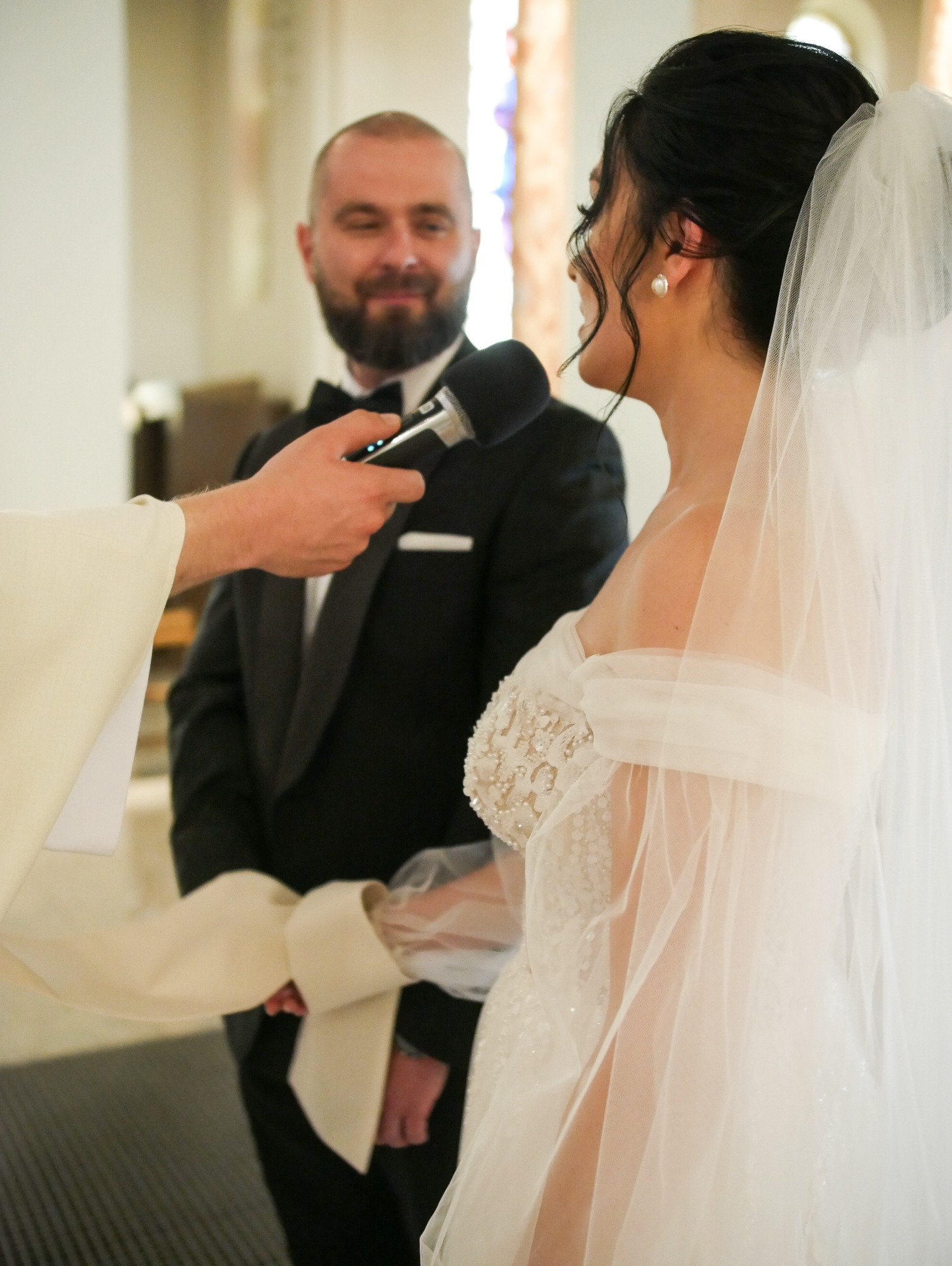 Uśmiechnięty pan młody w tle, podczas ceremonii ślubnej. W pierwszym planie fragment sukni panny młodej i dłoń trzymająca mikrofon.