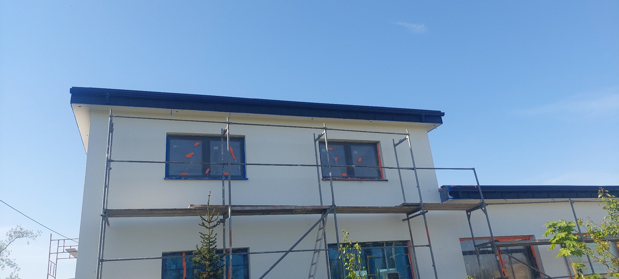 Biały dom z rusztowaniem, okna zabezpieczone folią. Widok na elewację w trakcie prac, błękitne niebo w tle. Detal wykończenia budynku.