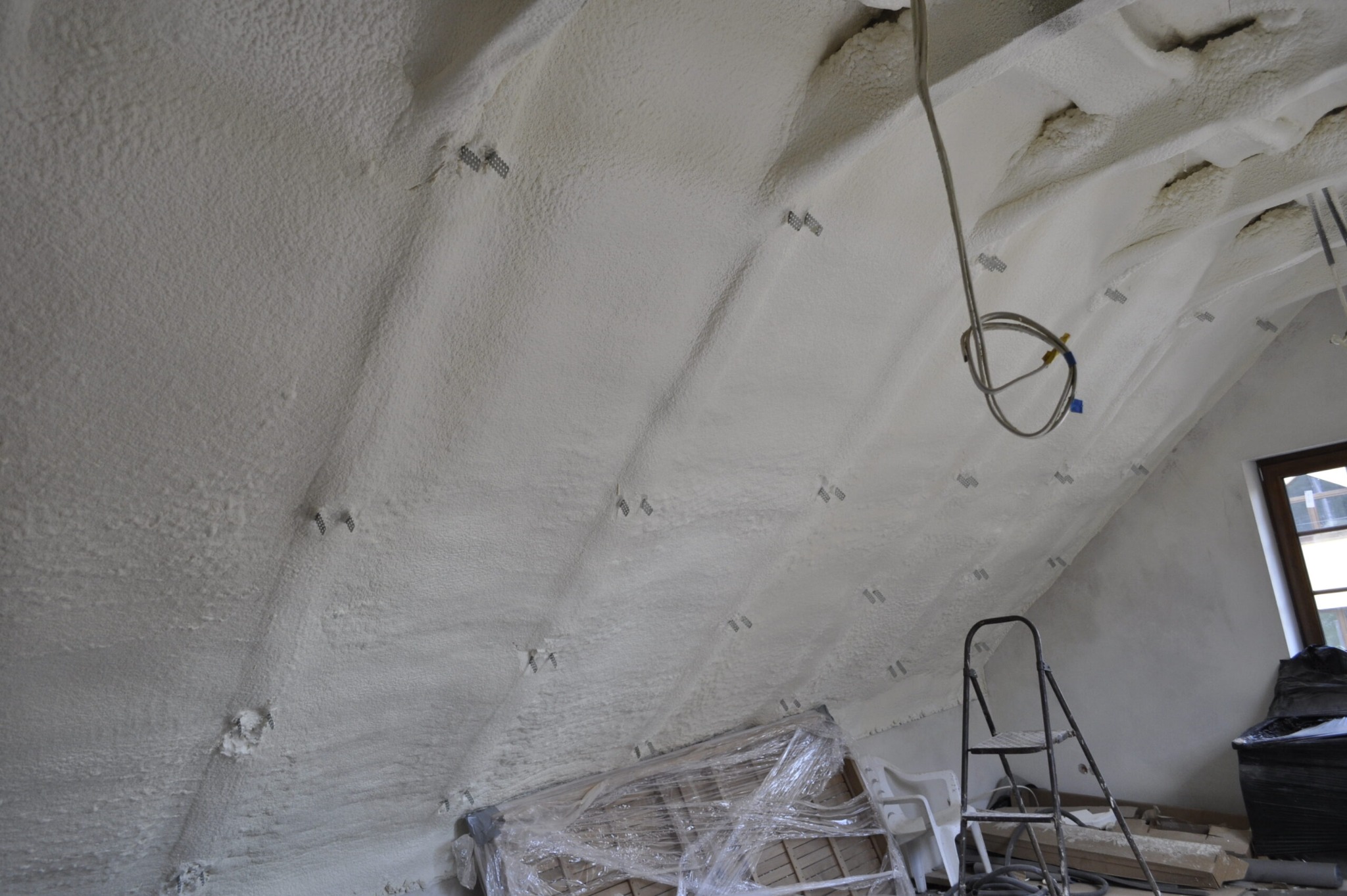 Poddasze po natrysku pianki, widoczne metalowe mocowania, przewody i drabina. Tekstura pianki nierówna, z widocznymi wgłębieniami. Okno w tle.