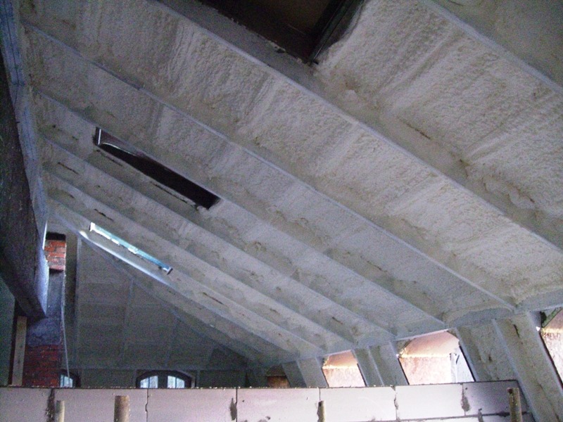 Poddasze z widoczną izolacją natryskową, wykonaną na skośnych belkach stropowych, z fragmentami okien dachowych. Widoczna ceglana ściana i fragment muru.